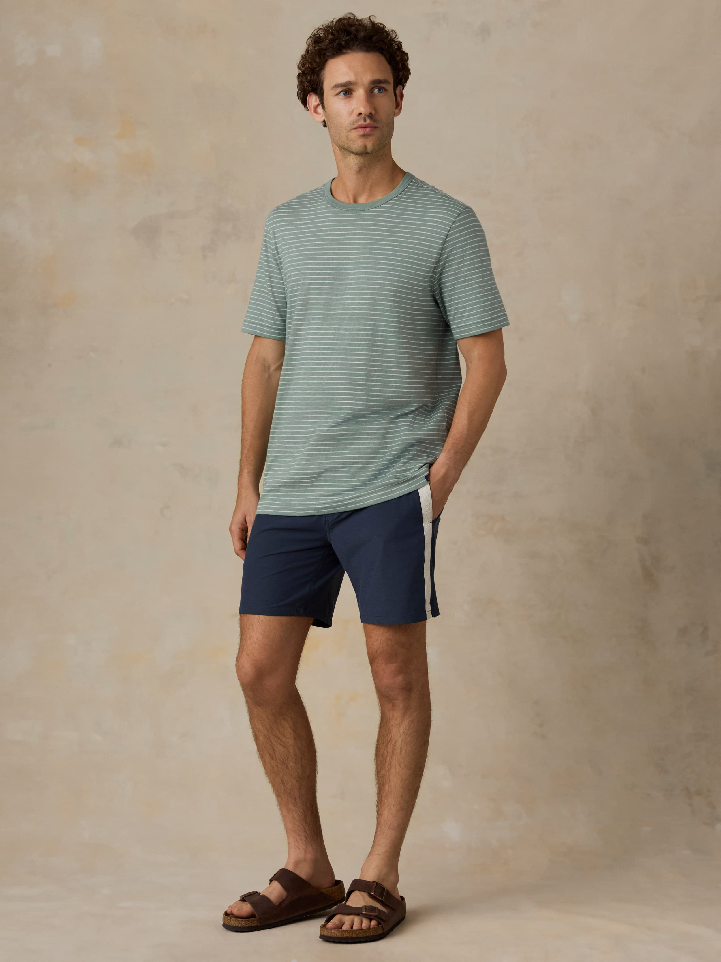 Man wearing a green and white striped tee with navy swim shorts against a beige background