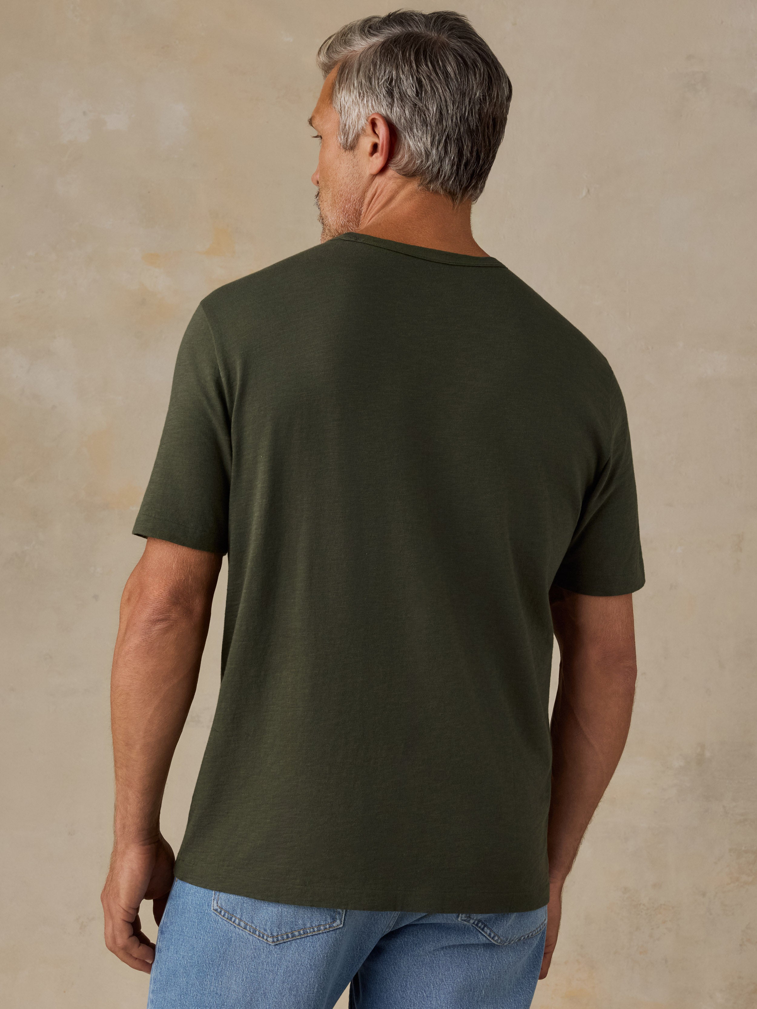 A man viewed from the back wearing a solid olive green short-sleeve t-shirt and blue jeans, standing against a textured beige studio wall.