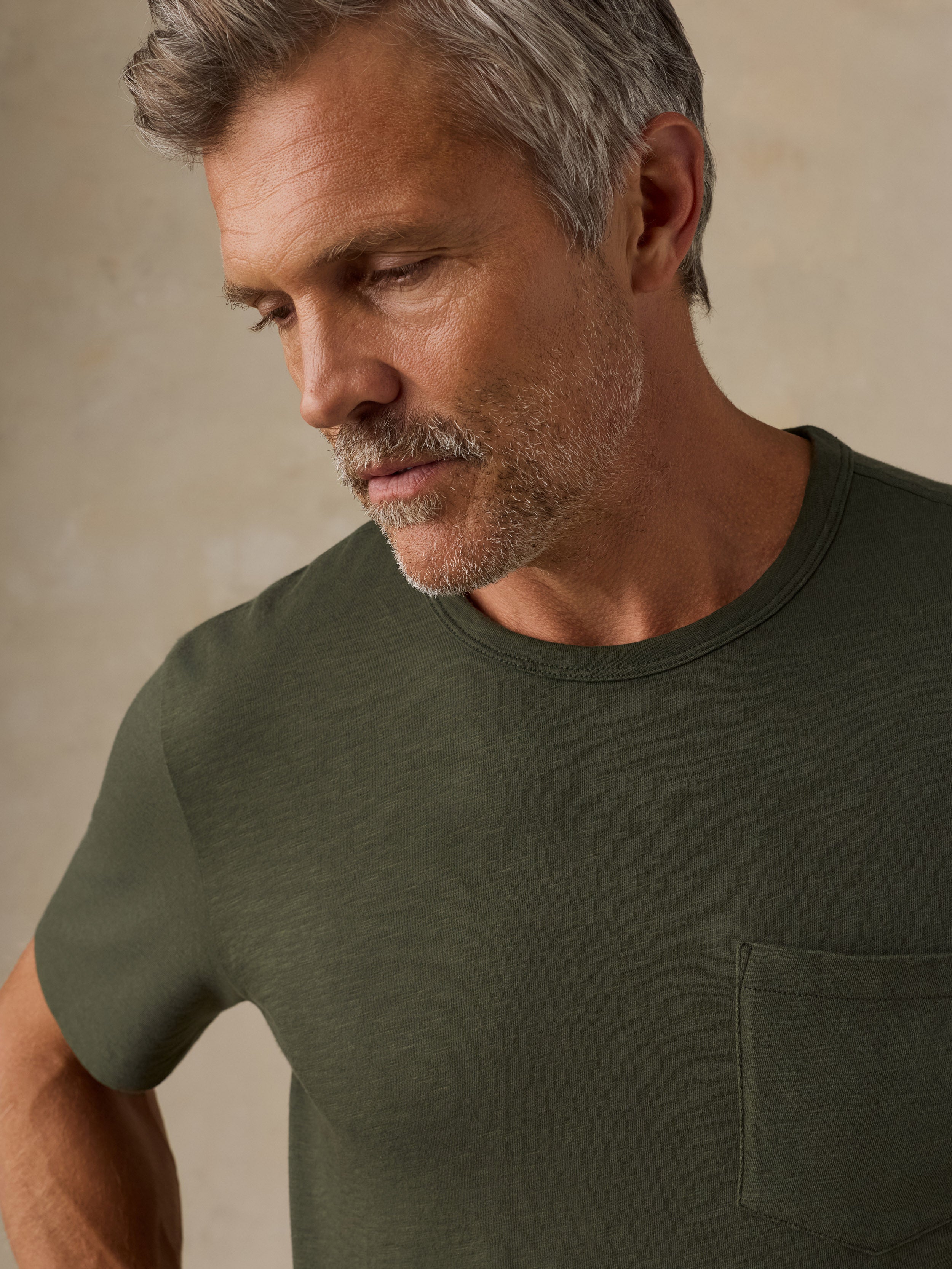 A close-up of a man wearing a dark olive green crew-neck t-shirt with a chest pocket, set against a neutral beige background.