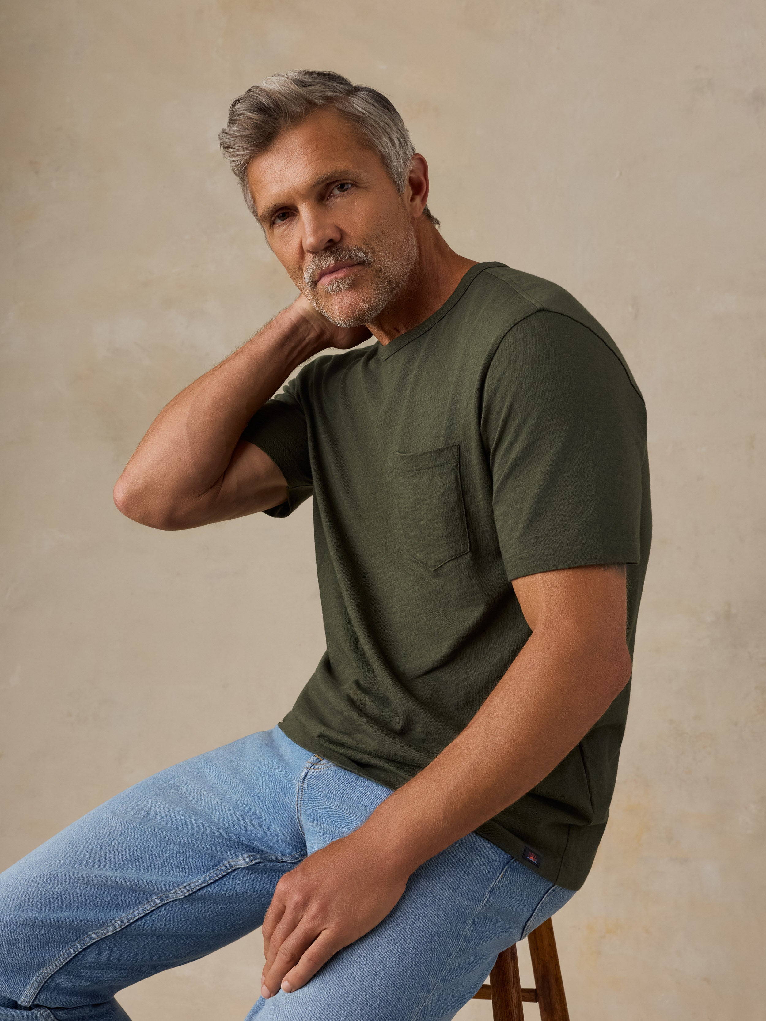 A man with gray hair and a beard sits on a wooden stool wearing an olive green short-sleeve pocket t-shirt and light blue jeans against a textured beige background.
