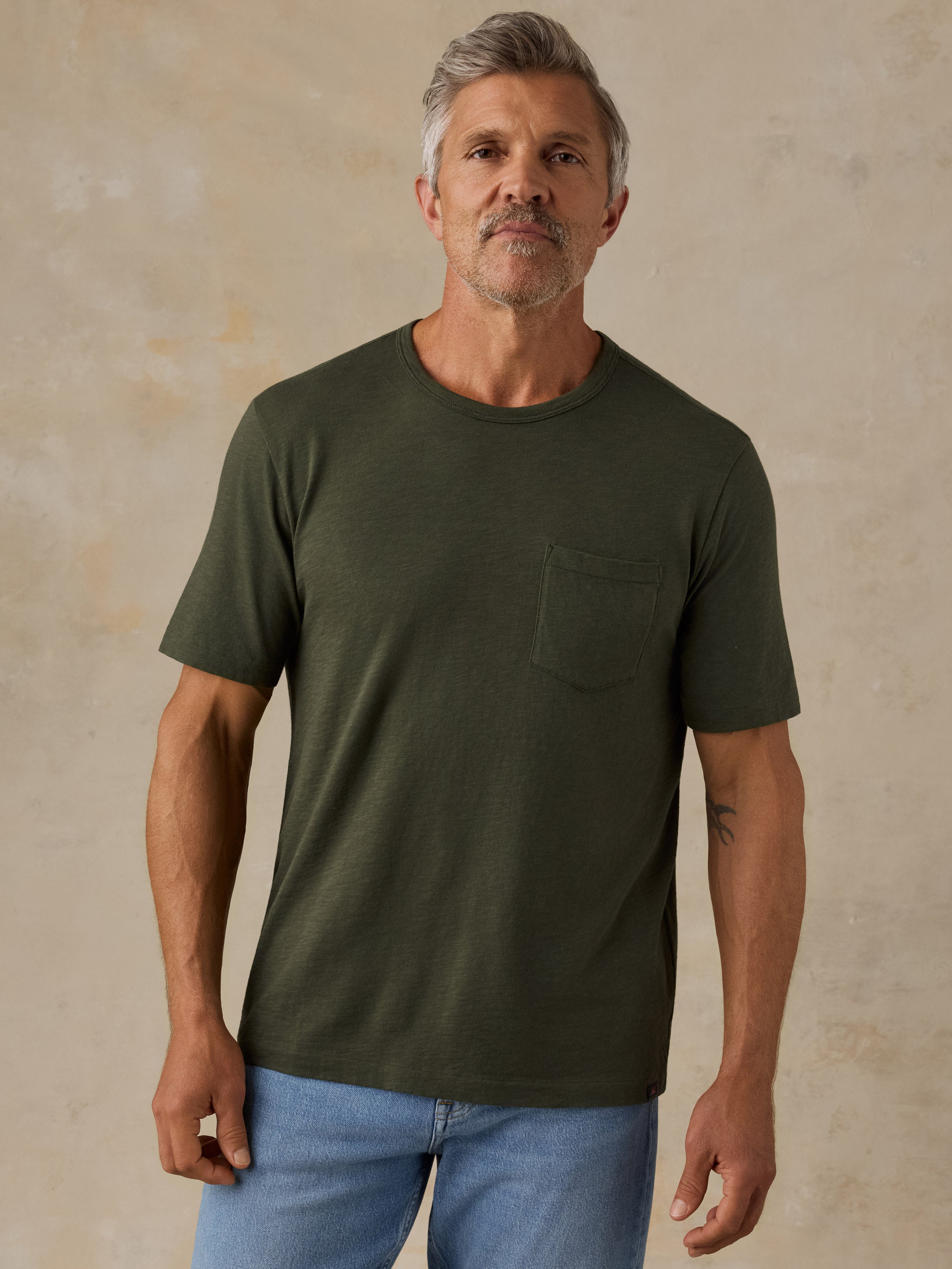A man wearing a dark olive green short-sleeve crewneck t-shirt with a chest pocket, paired with light blue jeans, standing against a textured beige wall.