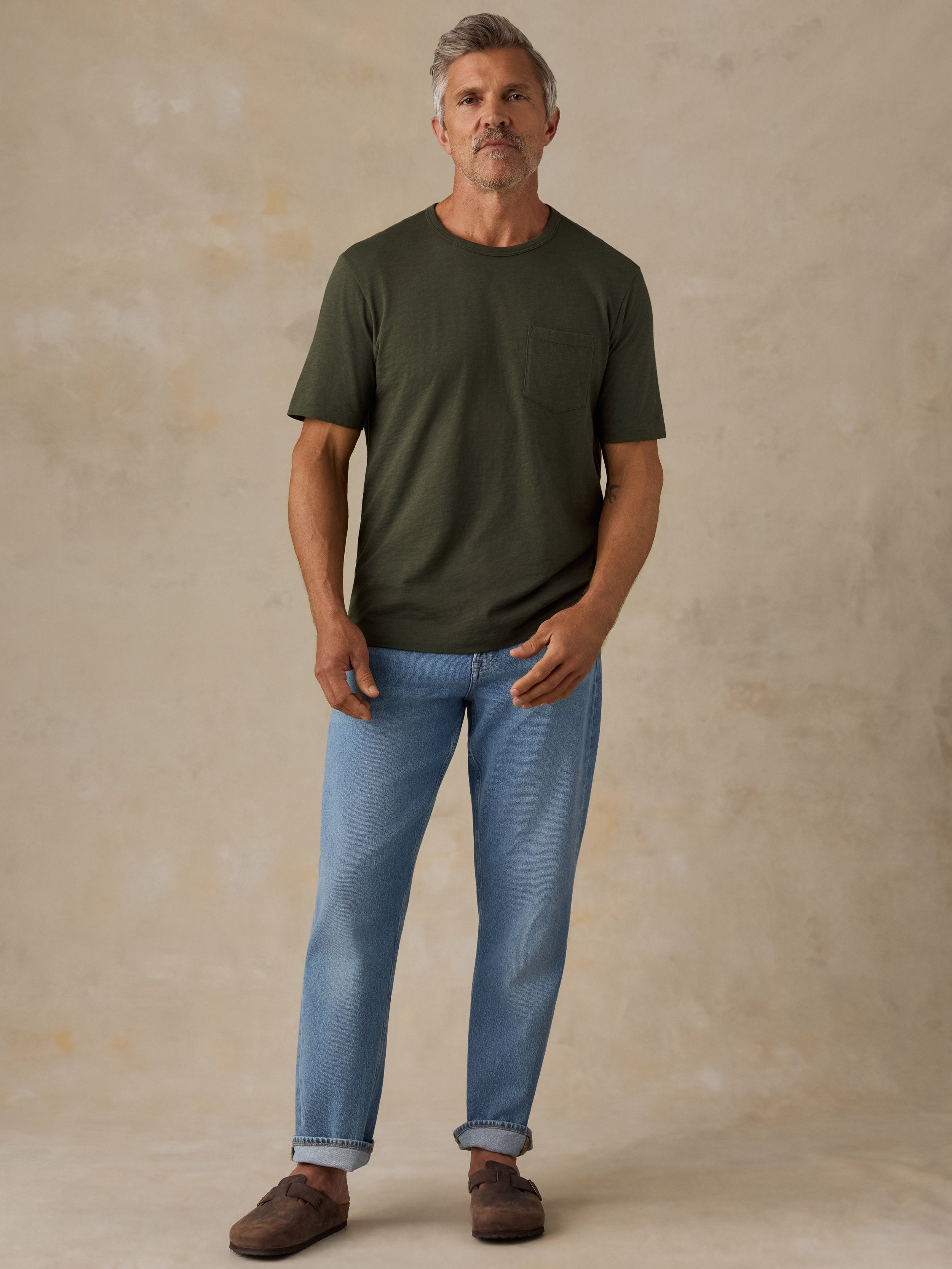 A man wearing an olive green short-sleeve crewneck pocket t-shirt, light blue jeans, and brown suede mules, standing against a textured beige studio backdrop.