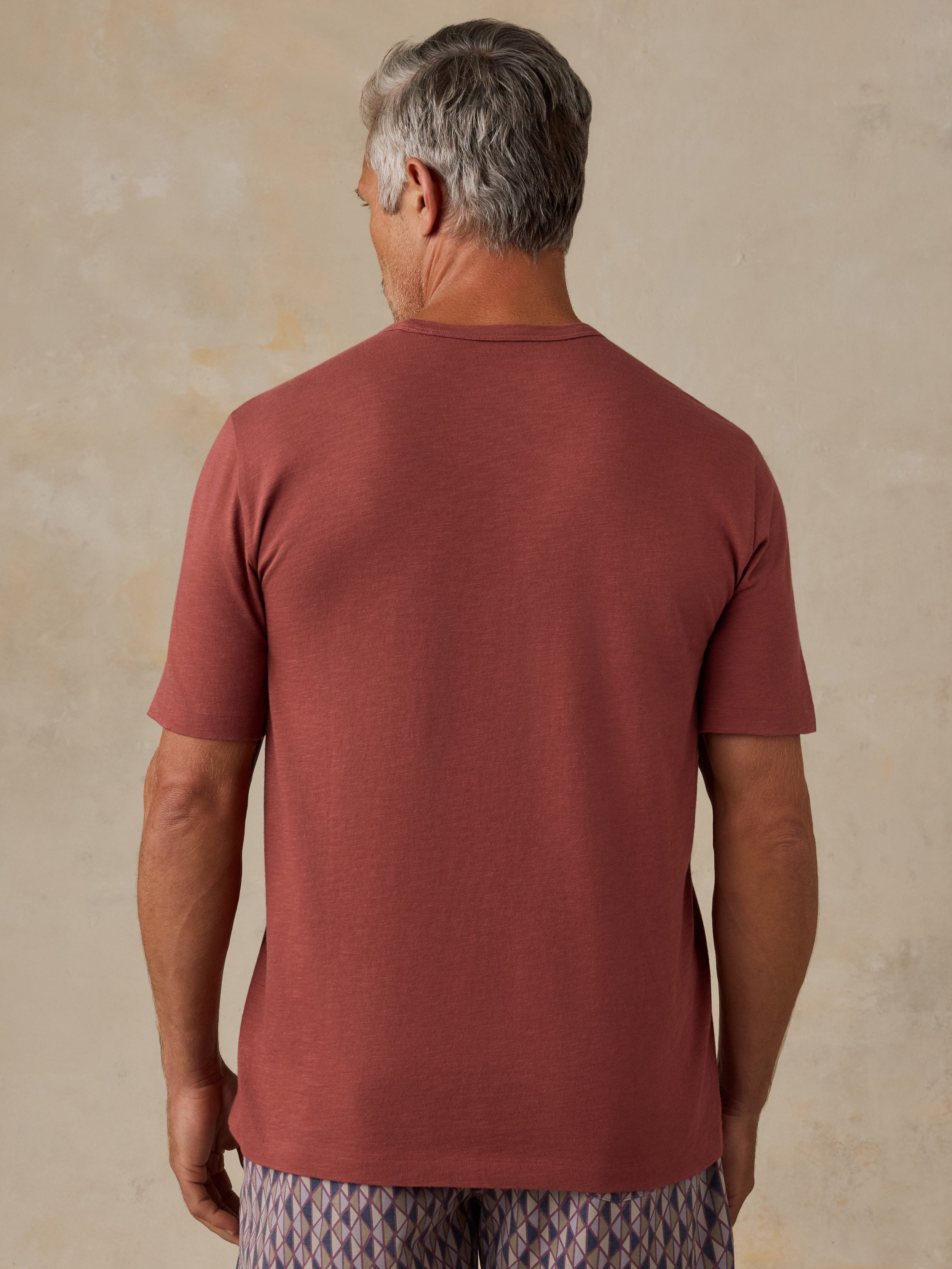 A man viewed from behind wearing a muted terracotta short-sleeve t-shirt and patterned trousers, standing against a textured beige wall.