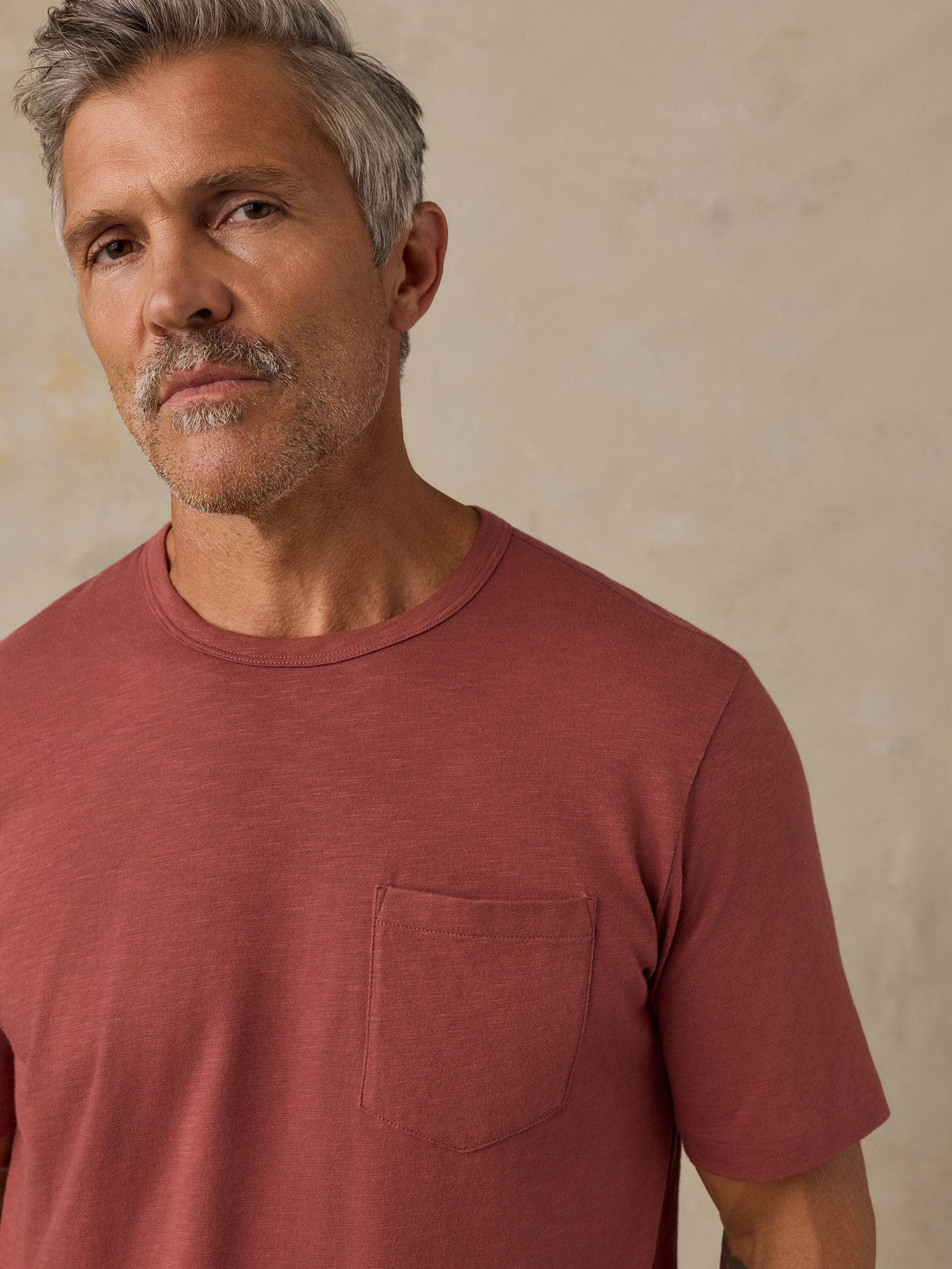 A close-up shot of a man wearing a muted terracotta crewneck t-shirt with a chest pocket, set against a textured beige wall.