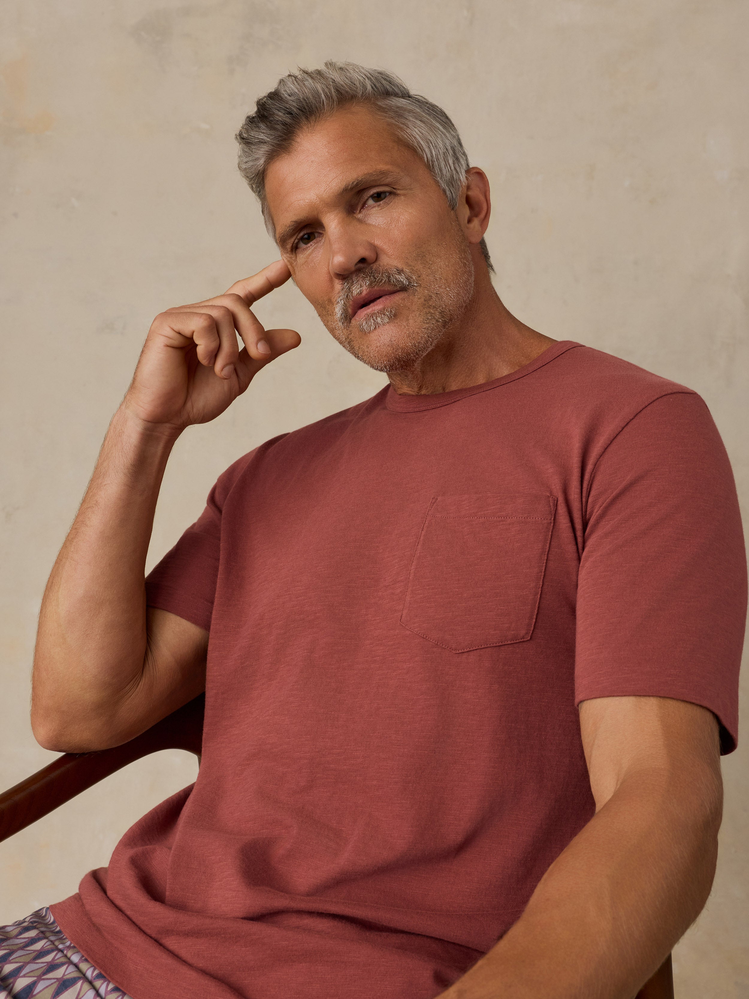 A man with gray hair and a mustache wearing a terracotta short-sleeve pocket t-shirt, sitting in a wooden chair against a textured beige wall.