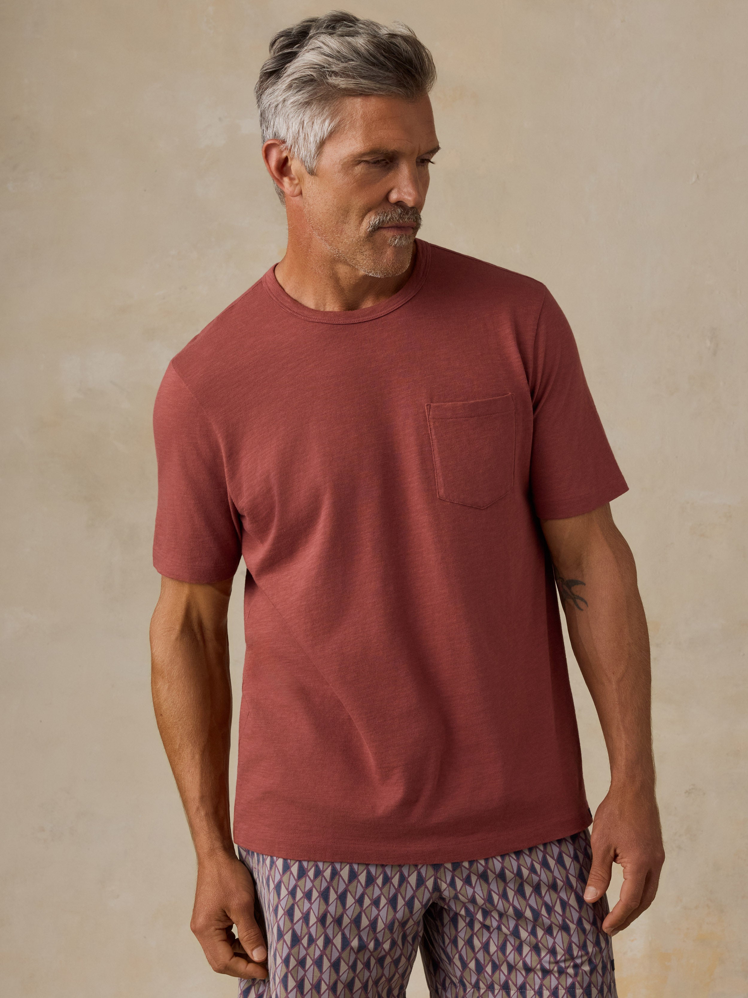 A man wearing a muted terracotta short-sleeve crewneck pocket t-shirt and patterned pants, standing against a textured beige wall.