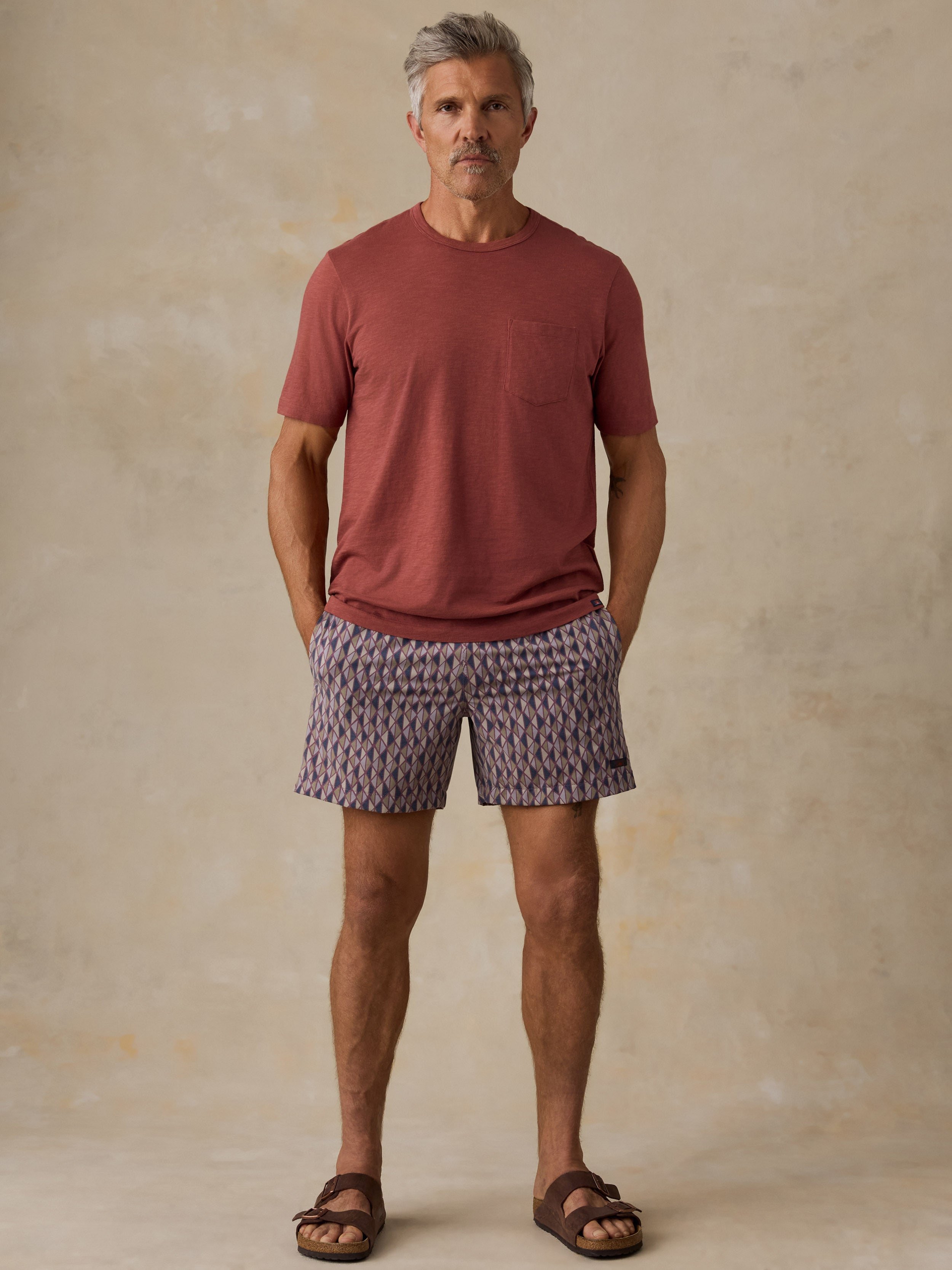 A man wearing a terracotta short-sleeve pocket t-shirt, patterned purple and beige shorts, and brown sandals, standing against a textured beige studio backdrop.