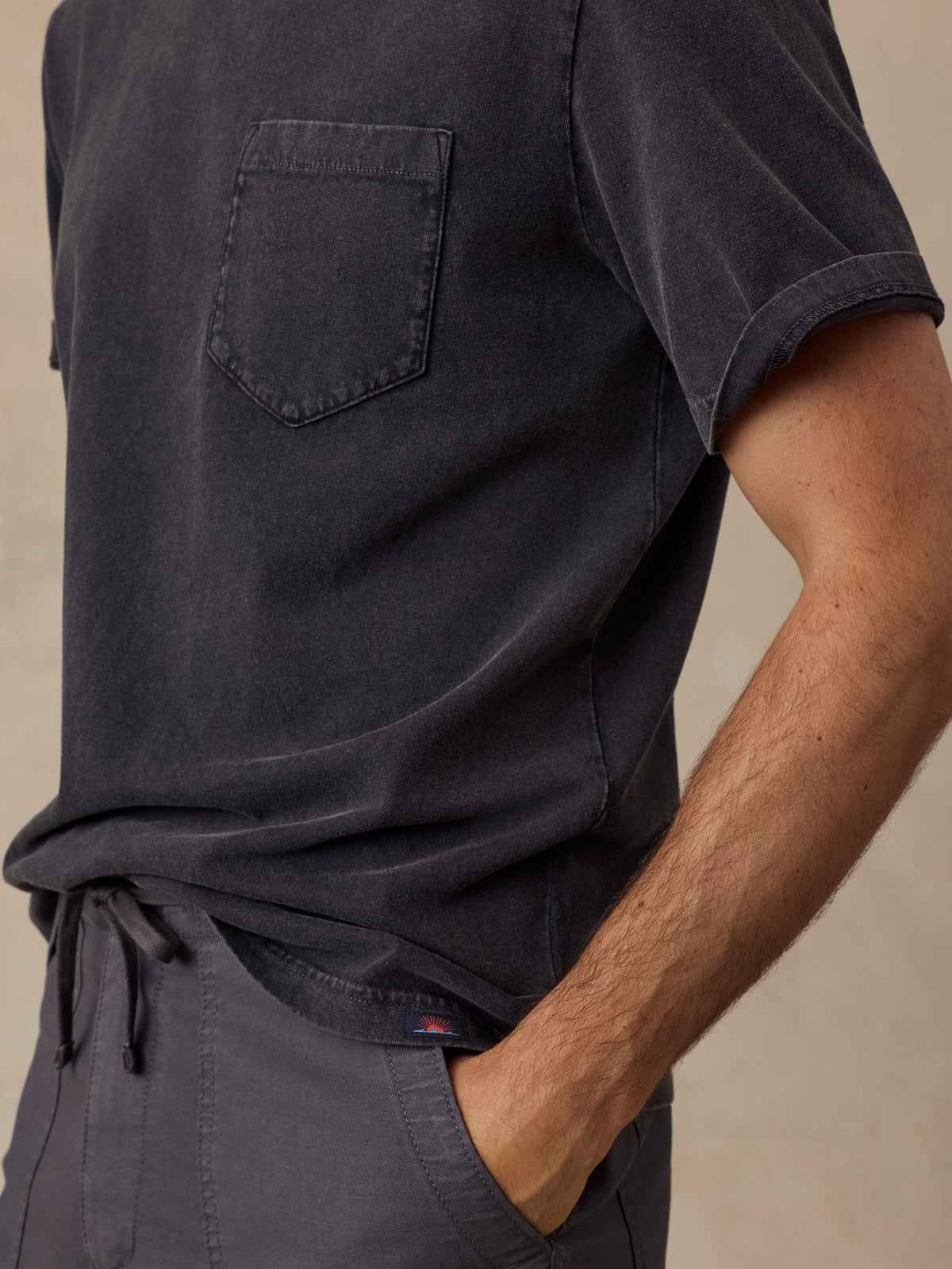 Close-up of a man wearing a dark charcoal gray short-sleeve t-shirt with a chest pocket and matching dark drawstring pants, against a neutral beige background., image 4 of 6