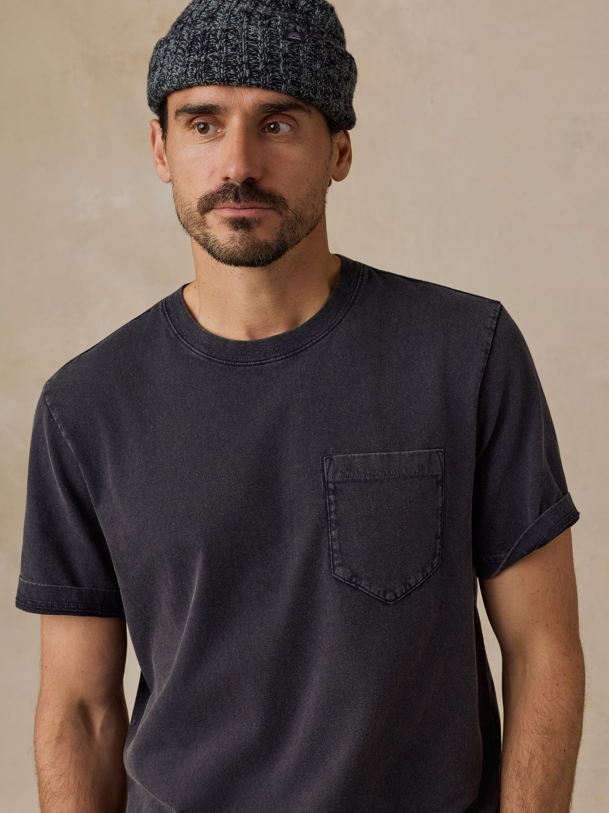 A man wearing a dark charcoal gray washed short-sleeve t-shirt with a chest pocket and a dark gray knit beanie, set against a neutral beige background., image 2 of 6