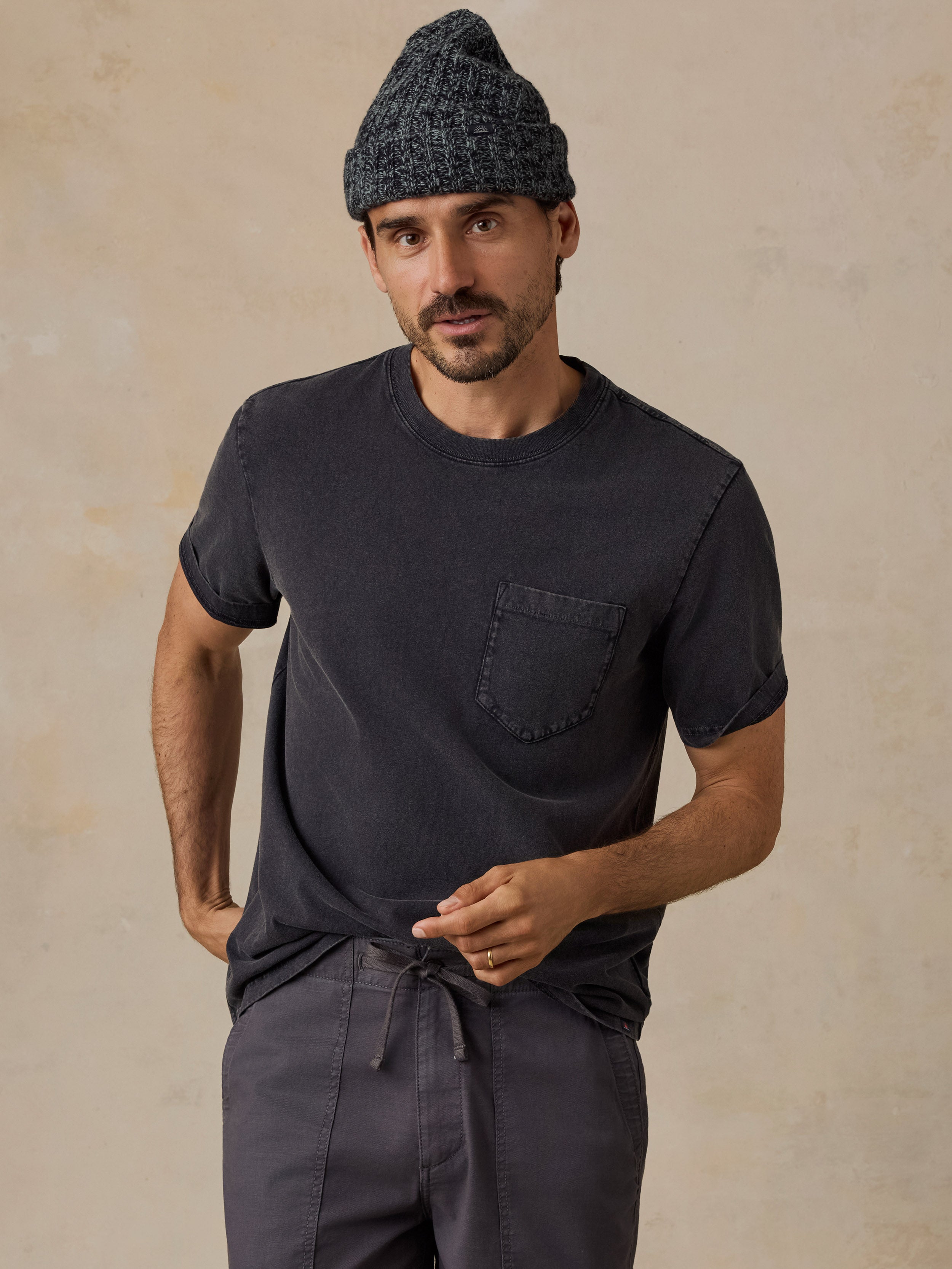 A man wearing a dark charcoal gray crewneck pocket t-shirt, matching dark gray drawstring pants, and a dark gray knit beanie, standing against a textured beige studio background.