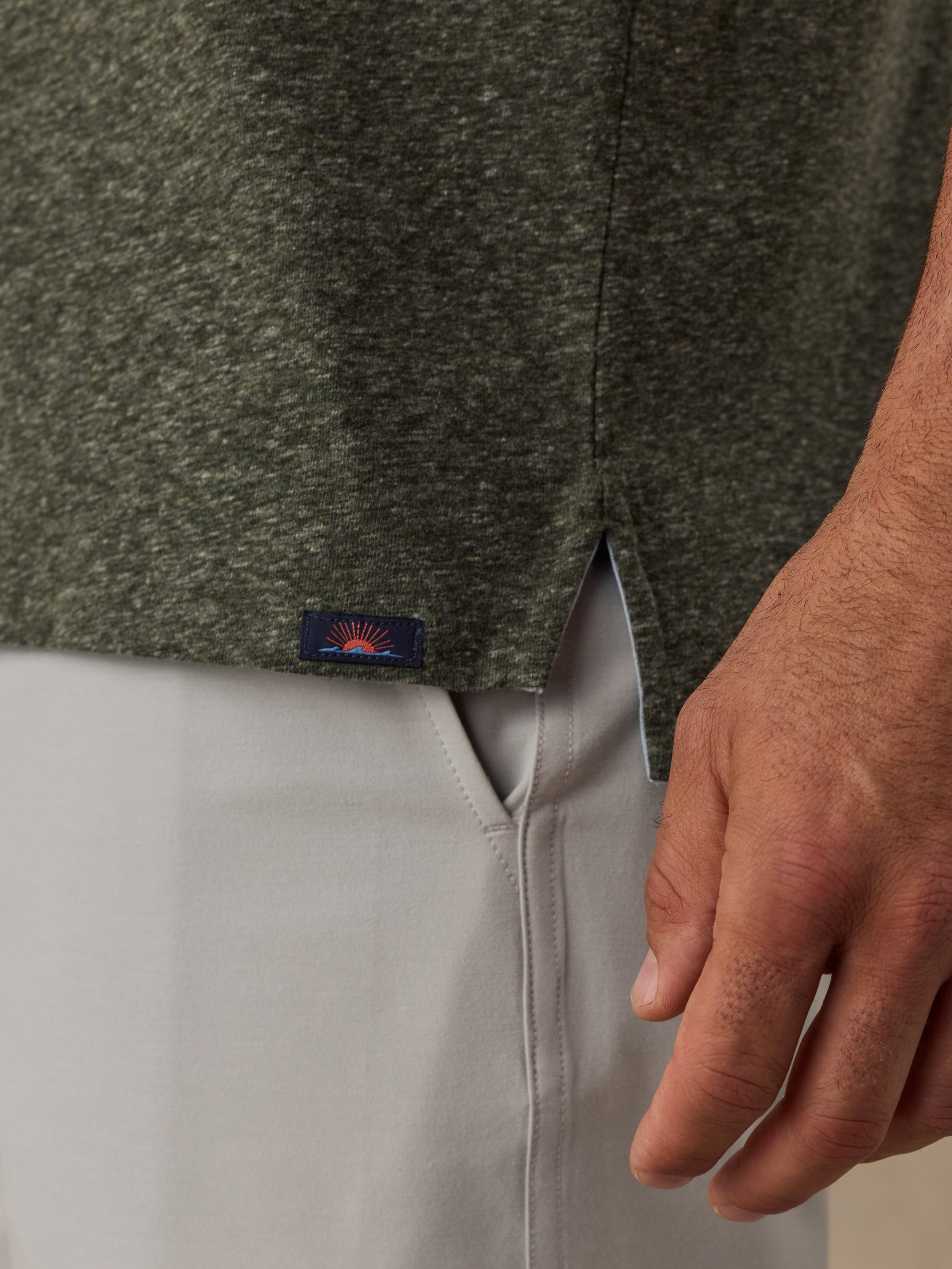 Close-up of a man wearing a dark heathered green knit top and light gray pants, with a small Faherty brand patch near the hem.