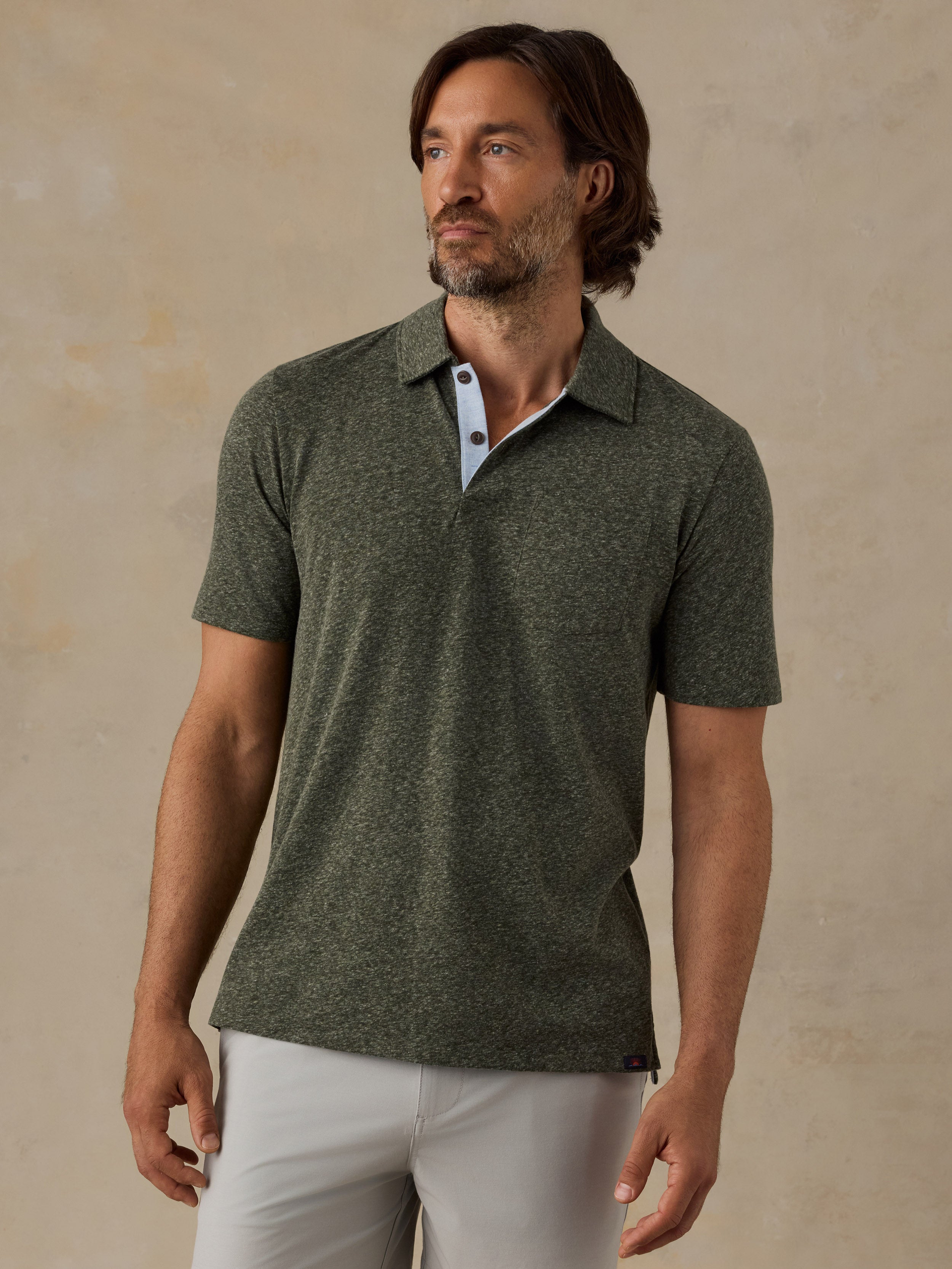 A man wearing a short-sleeve polo shirt in heathered olive green, paired with light-colored trousers, standing against a textured beige wall.