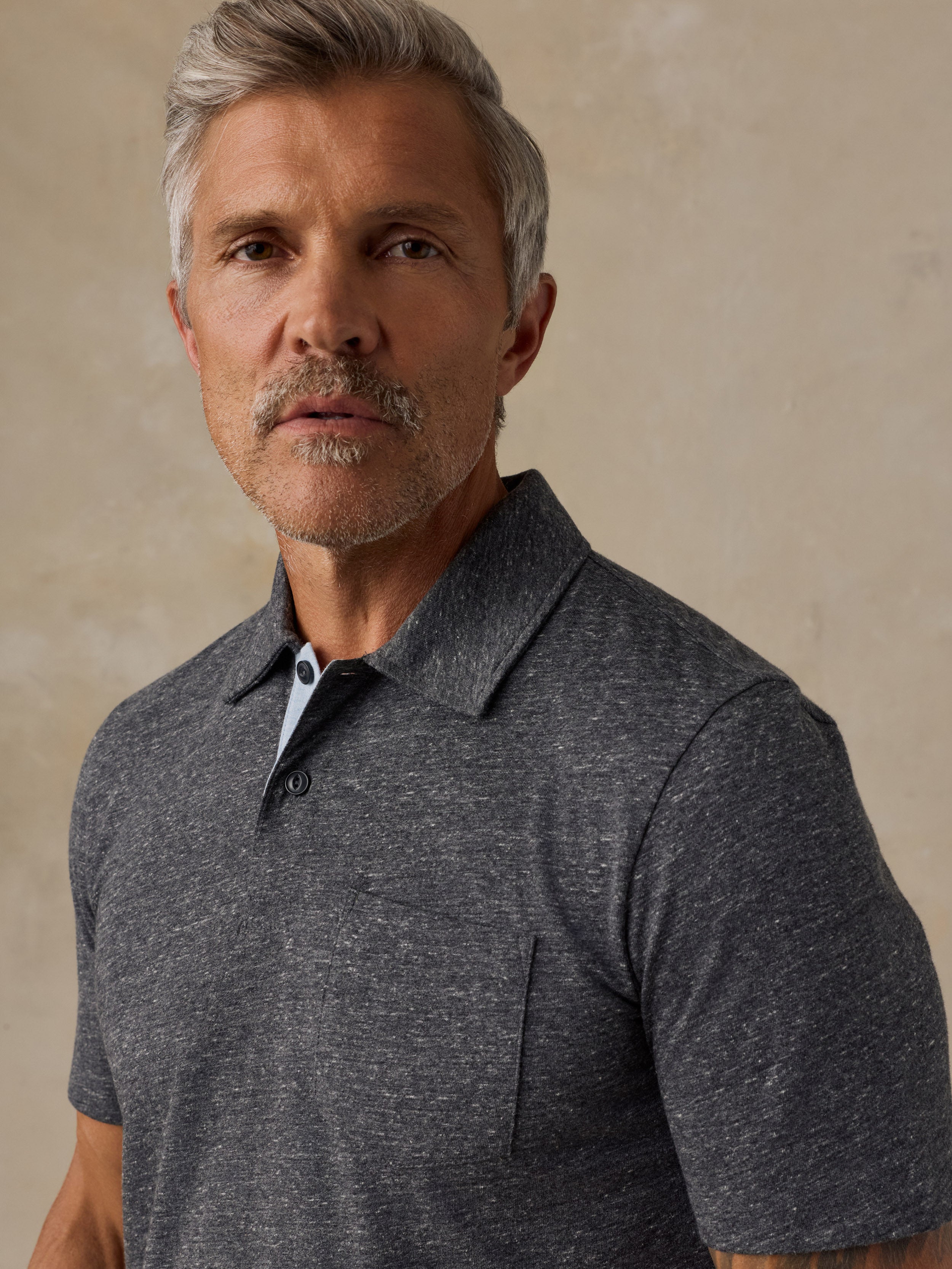 A close-up shot of a man wearing a dark heather gray short-sleeve polo shirt with a classic collar and a buttoned placket, set against a textured beige background.