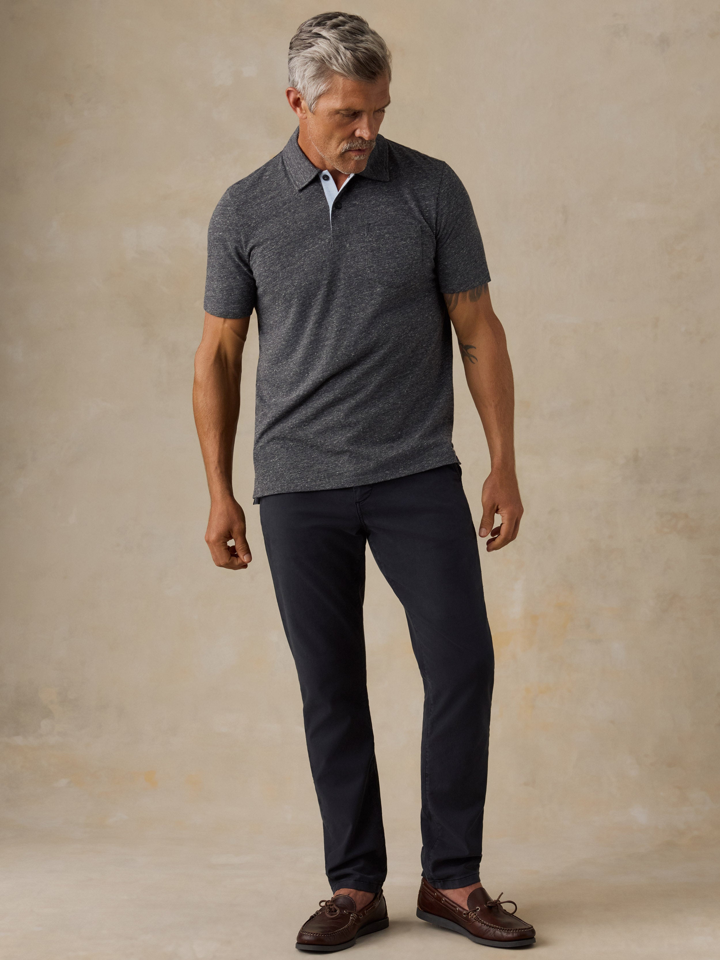 A man wearing a dark gray short-sleeve knit polo shirt, dark navy trousers, and brown boat shoes, standing against a textured beige studio backdrop.