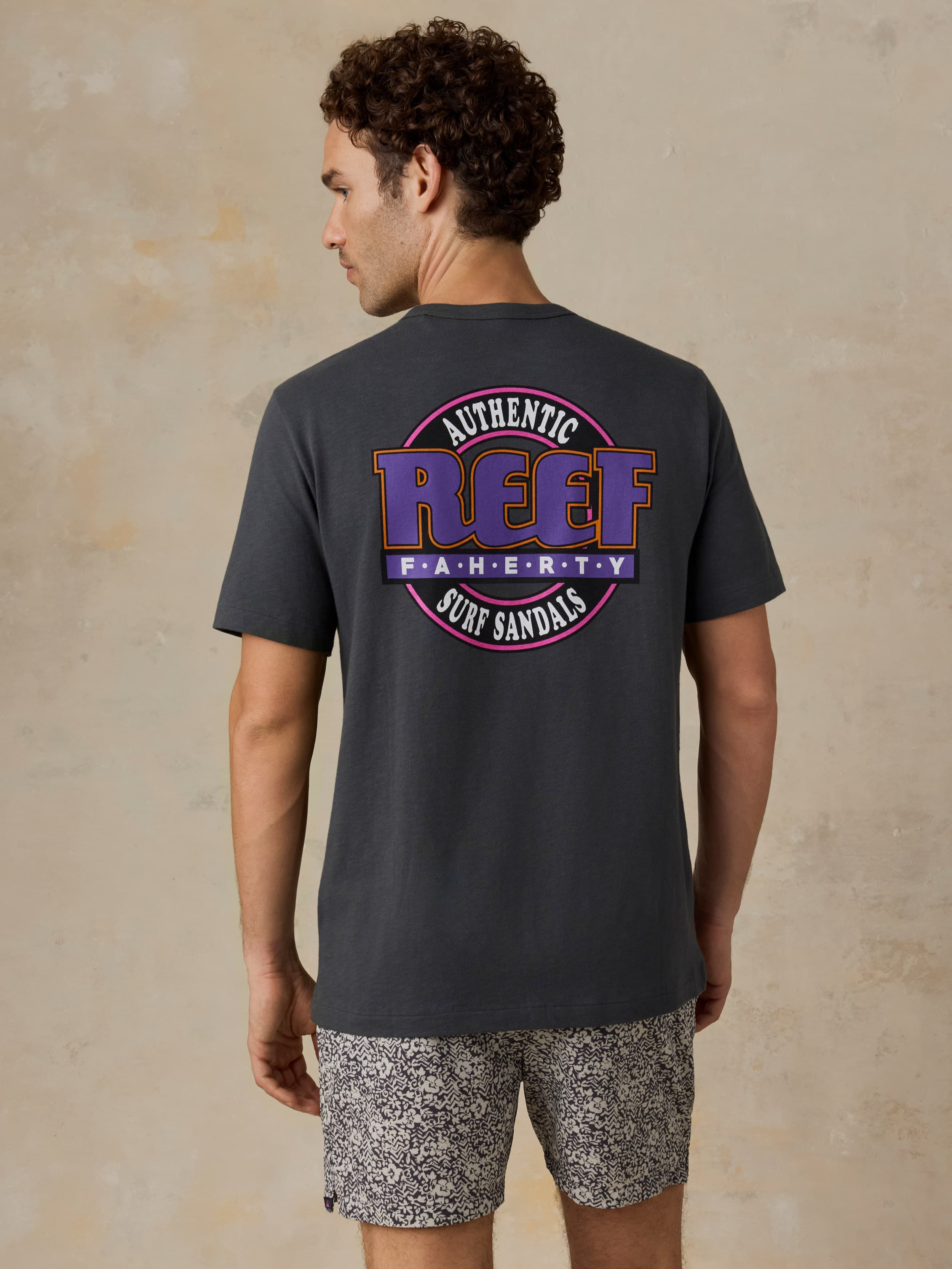 Man wearing a dark gray t-shirt with a Faherty x REEF logo and patterned shorts on a beige background