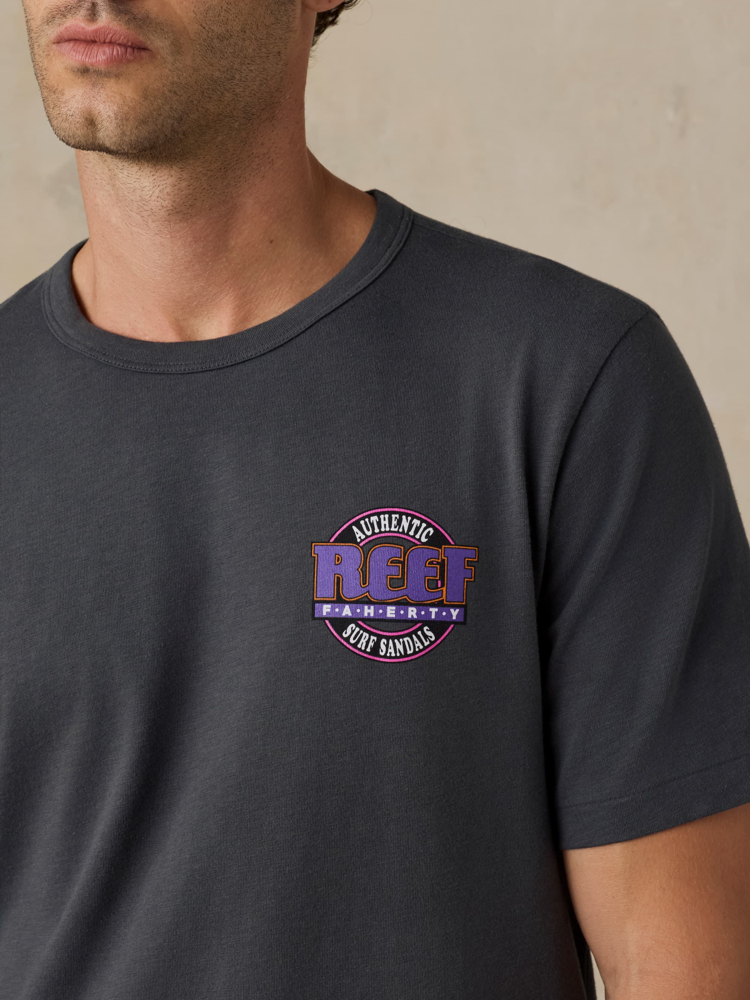 Man wearing a dark gray t-shirt with a Faherty x REEF logo on a beige background