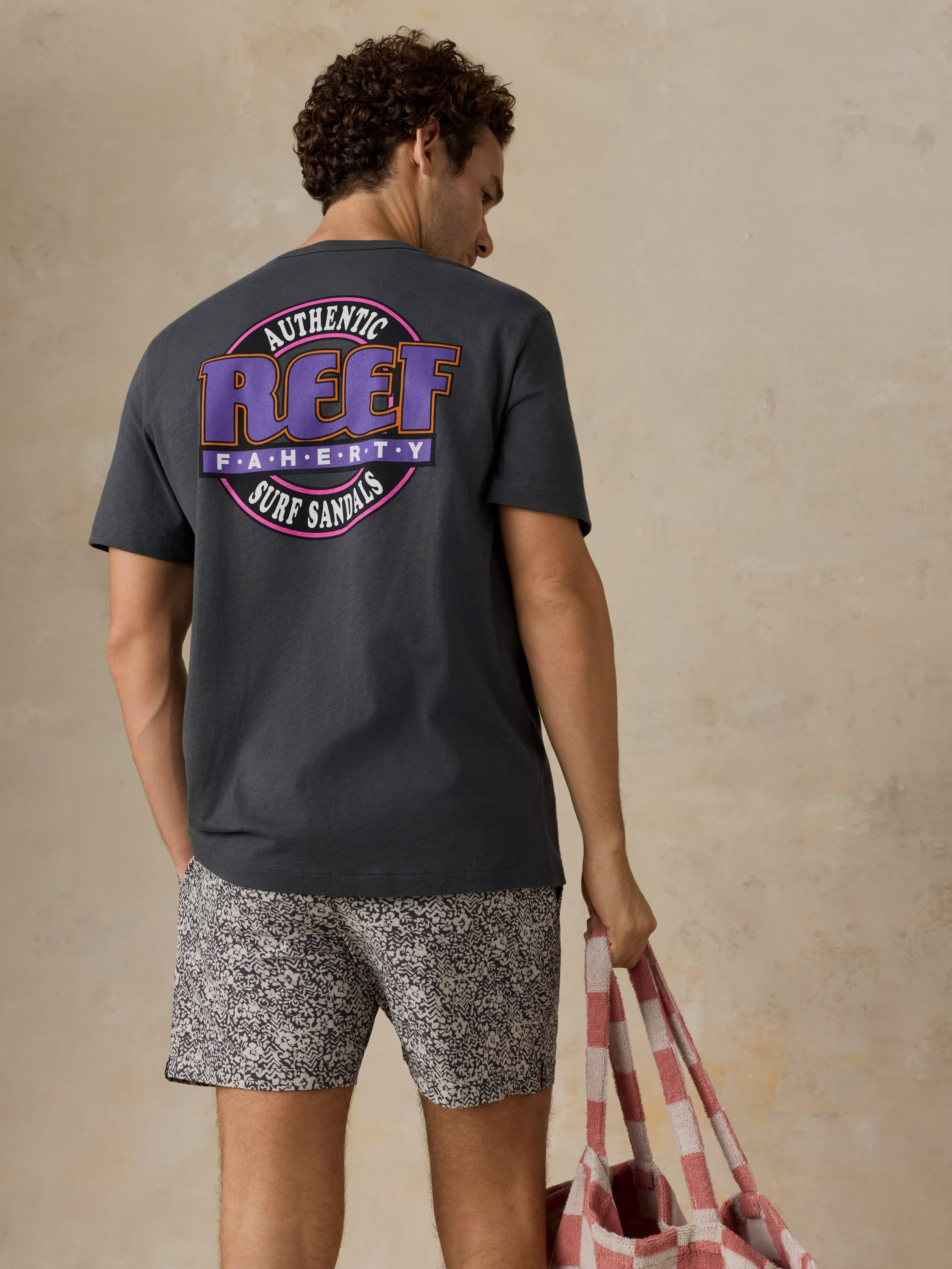 Man wearing a dark gray t-shirt with a Faherty x REEF logo and patterned shorts on a beige background