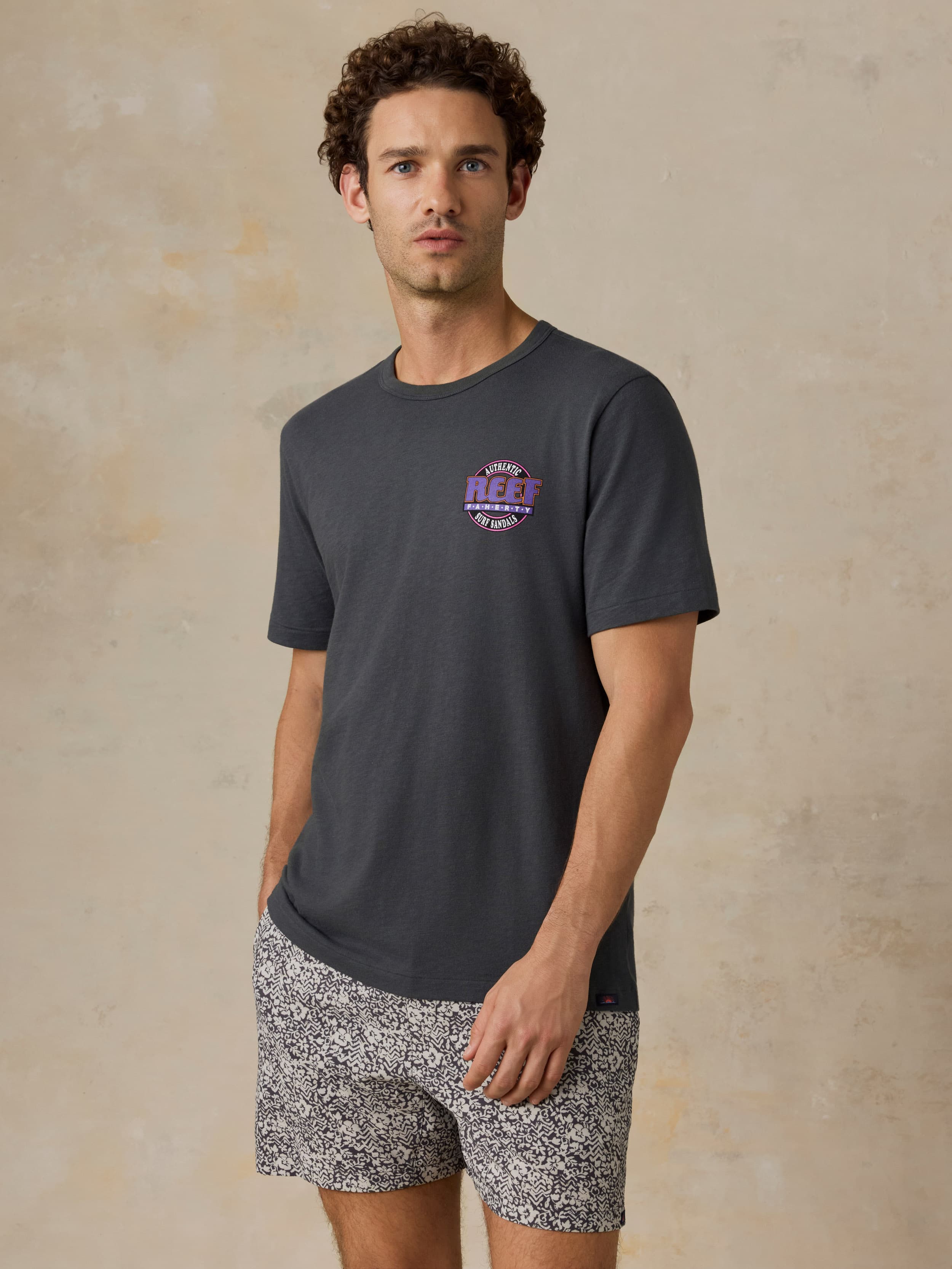 Man wearing a dark gray t-shirt with a Faherty x REEF logo and patterned shorts on a beige background
