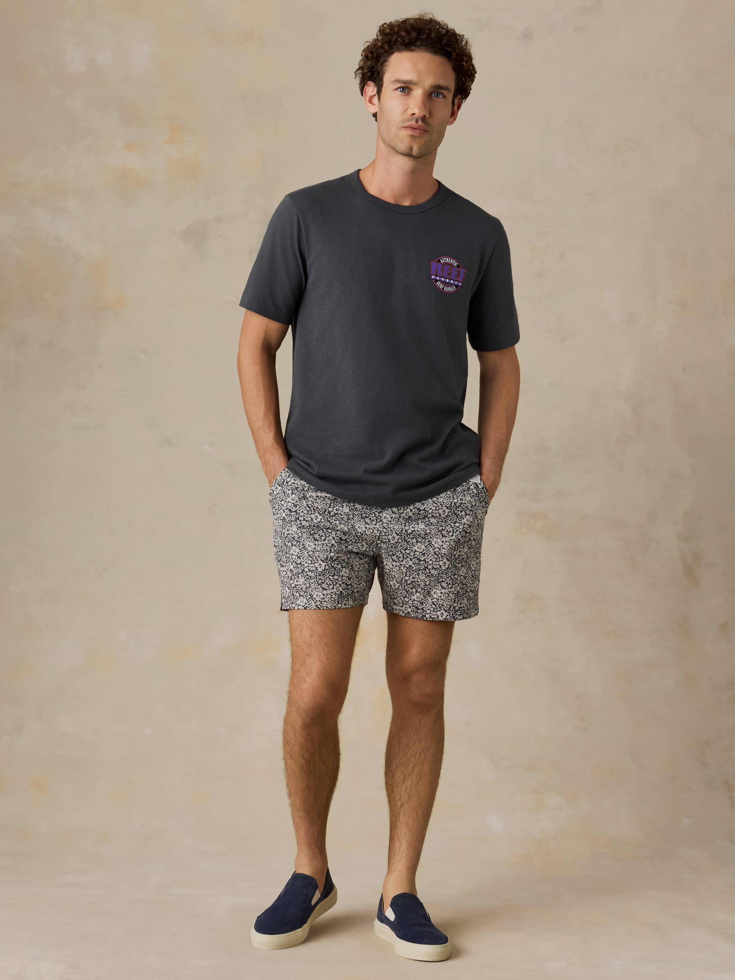 Man wearing a dark gray t-shirt with a Faherty x REEF logo and patterned shorts on a beige background