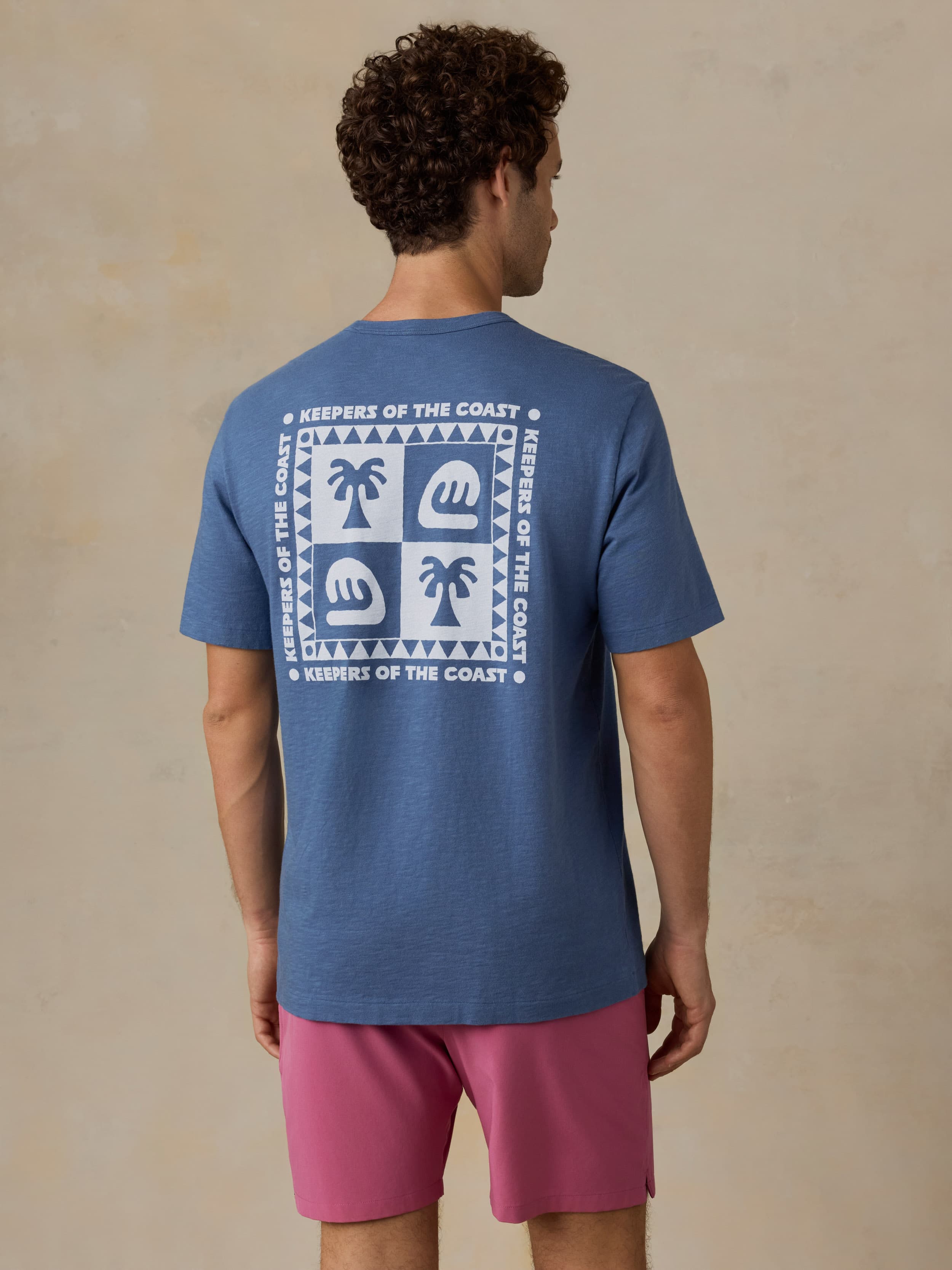 Man wearing a blue t-shirt with a Faherty x Surfrider Foundation logo and pink shorts against a beige background