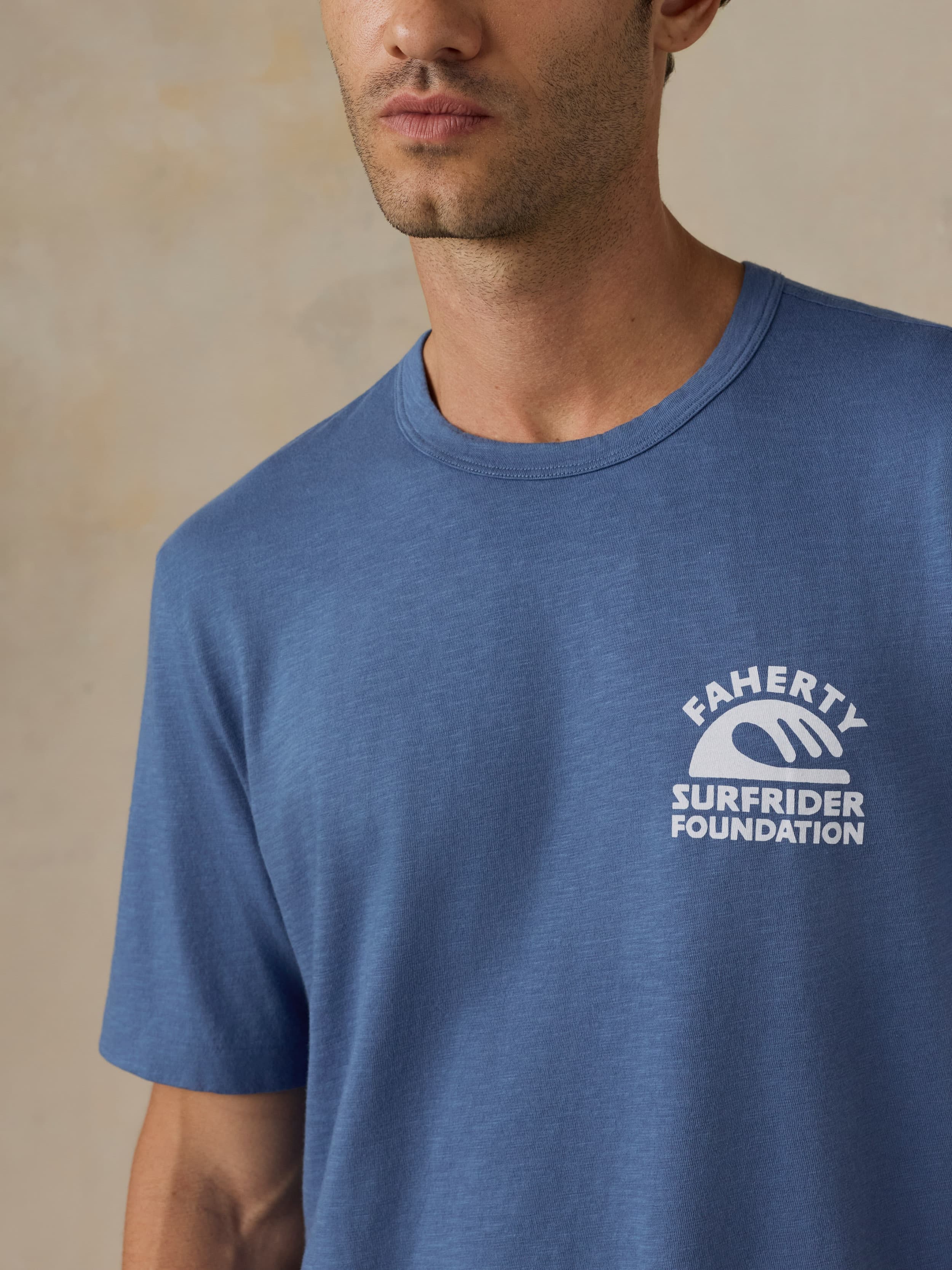 Man wearing a blue t-shirt with a Faherty x Surfrider Foundation logo against a beige background
