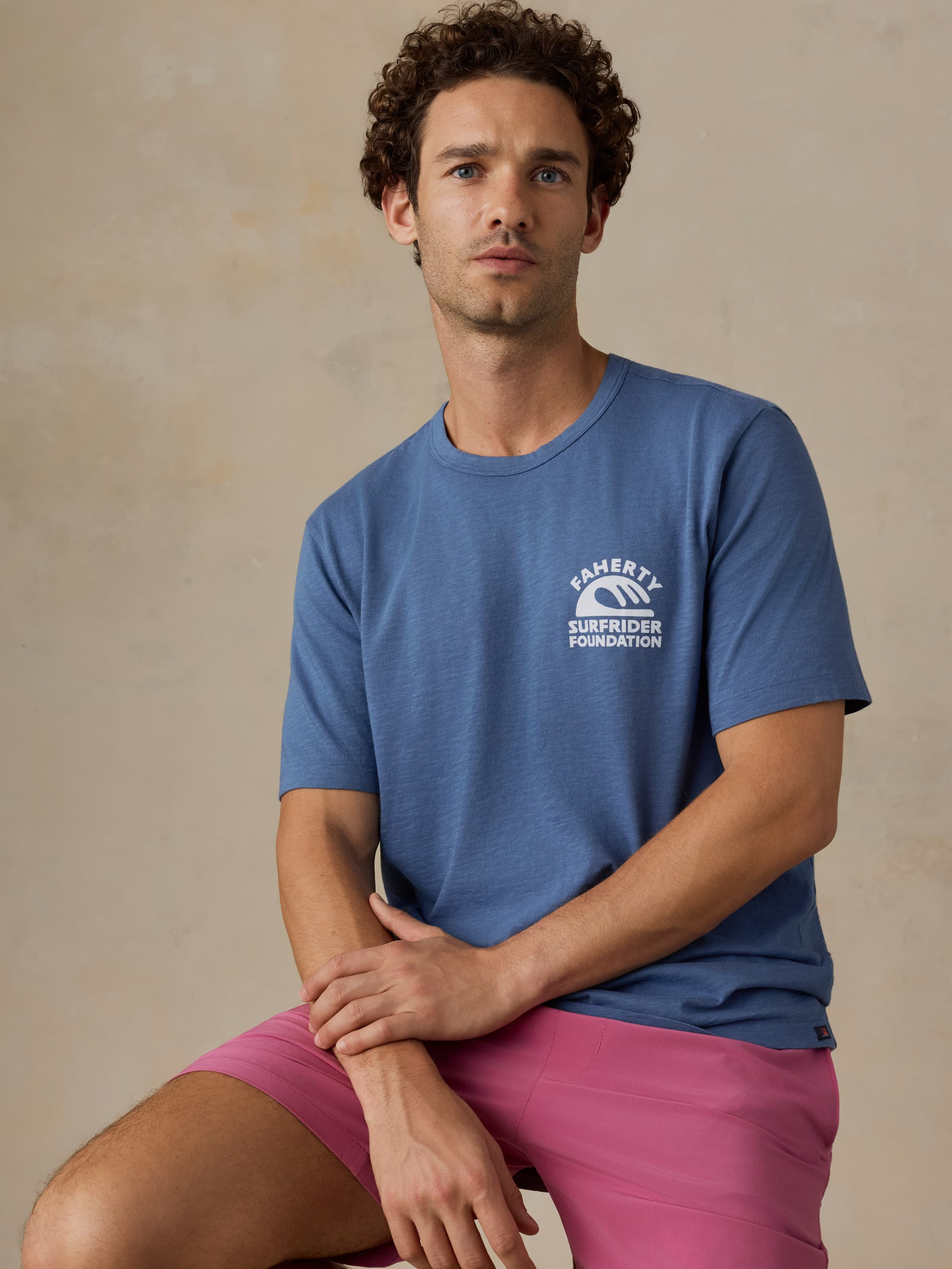Man wearing a blue t-shirt with a Faherty x Surfrider Foundation logo and pink shorts against a beige background