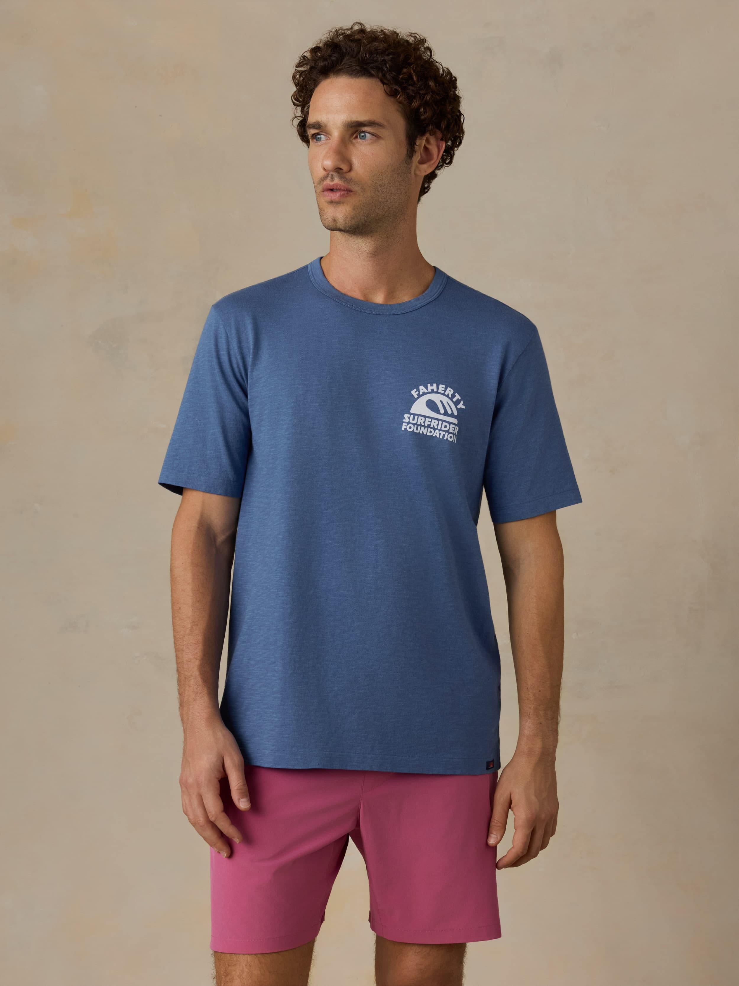 Man wearing a blue t-shirt with a Faherty x Surfrider Foundation logo and pink shorts against a beige background