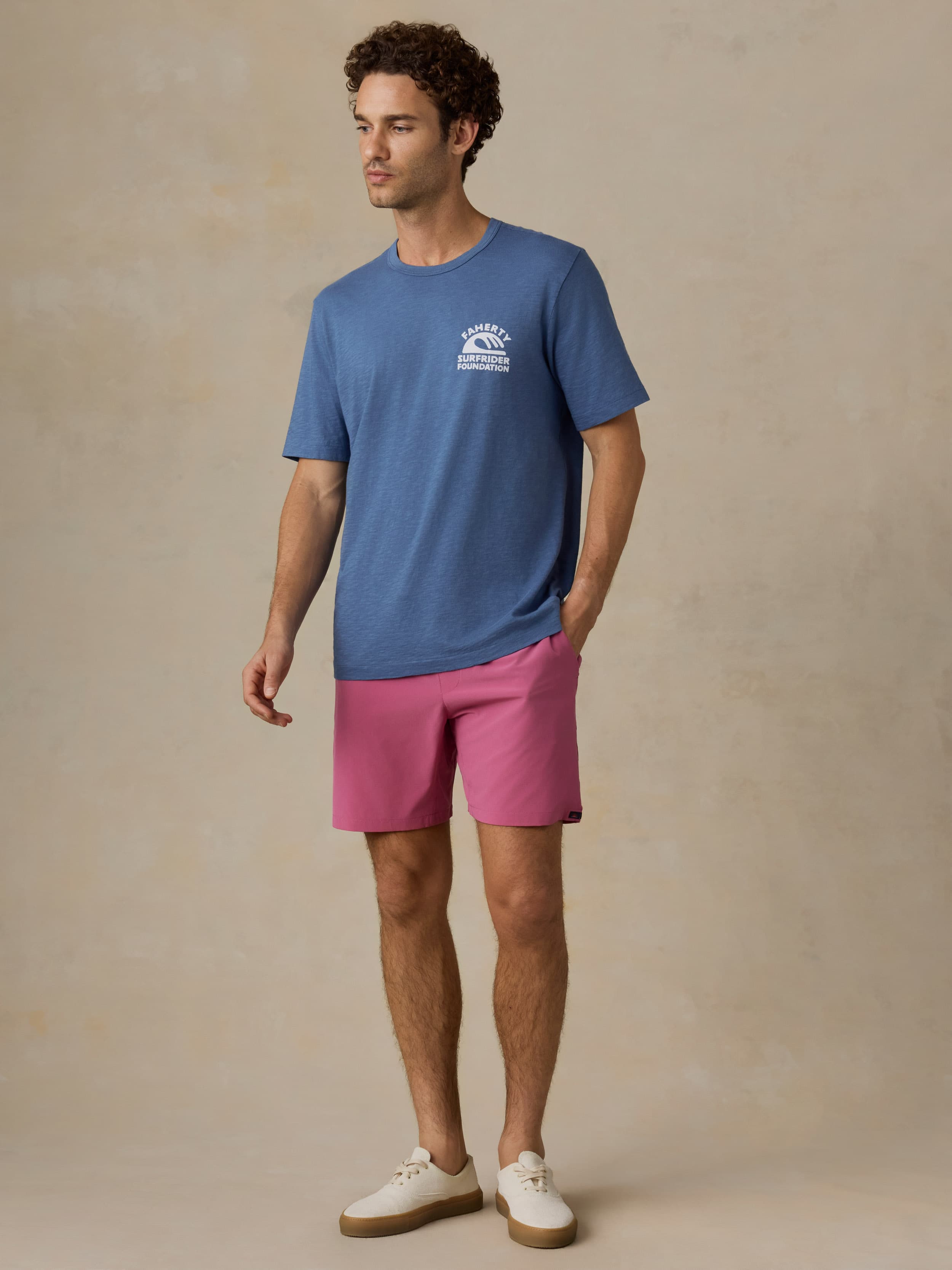Man wearing a blue t-shirt with a Faherty x Surfrider Foundation logo and pink shorts against a beige background