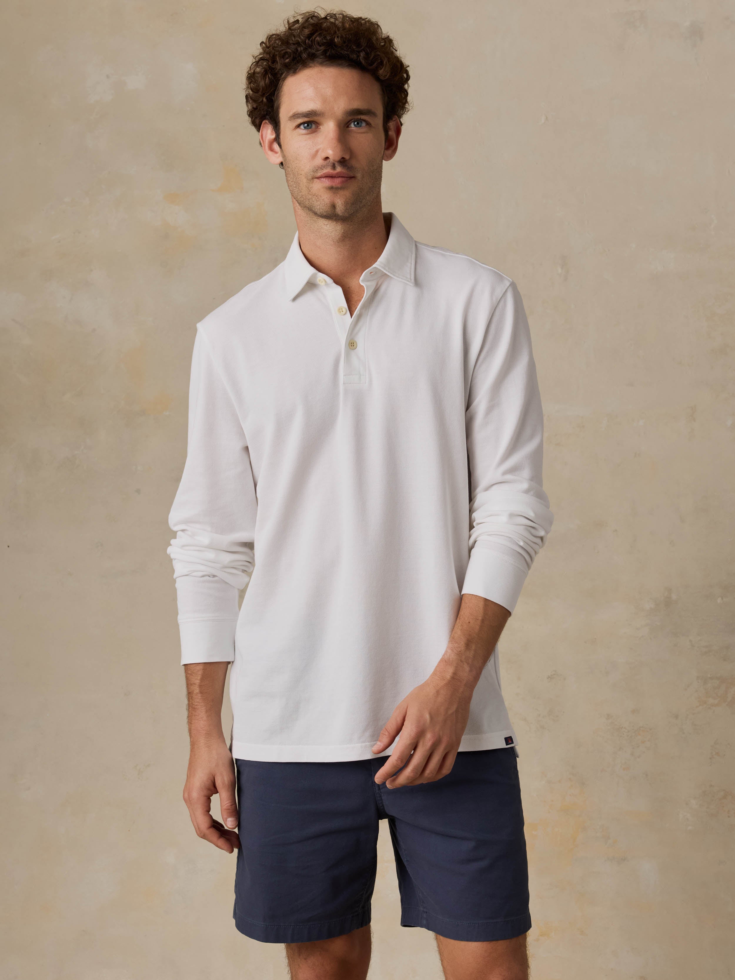 Man wearing a white long-sleeve polo shirt and navy shorts against a beige background