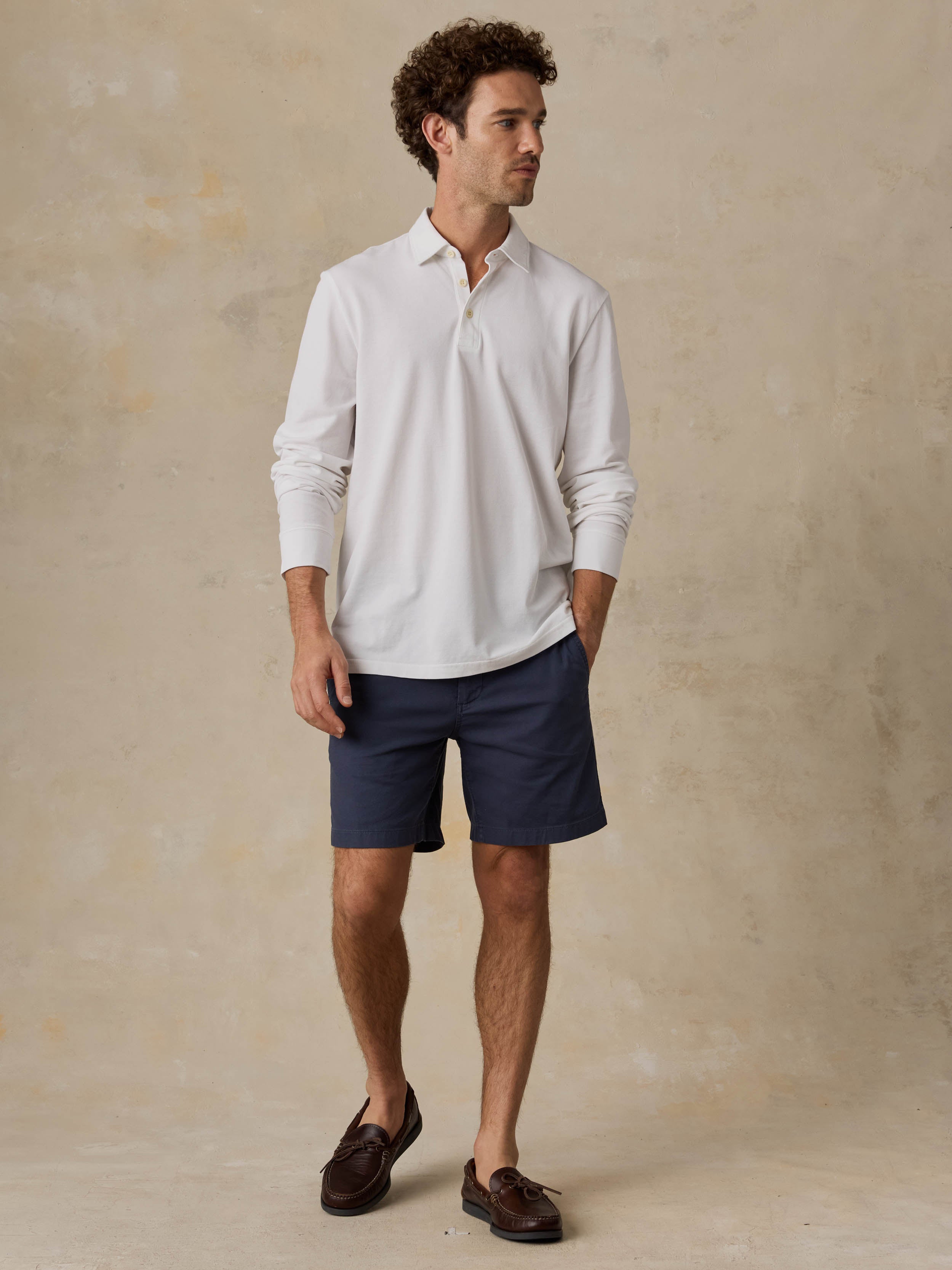 Man wearing a white long-sleeve polo shirt and navy shorts against a beige background