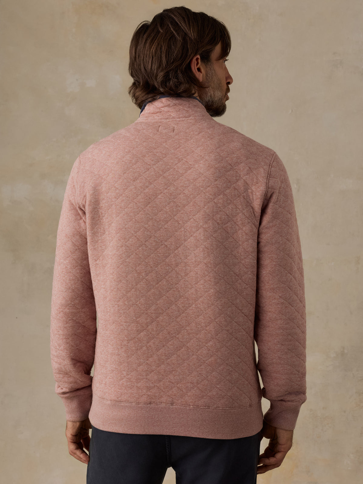 A man viewed from behind wearing a dusty rose quilted knit sweater and dark trousers, standing against a textured beige wall., image 2 of 5