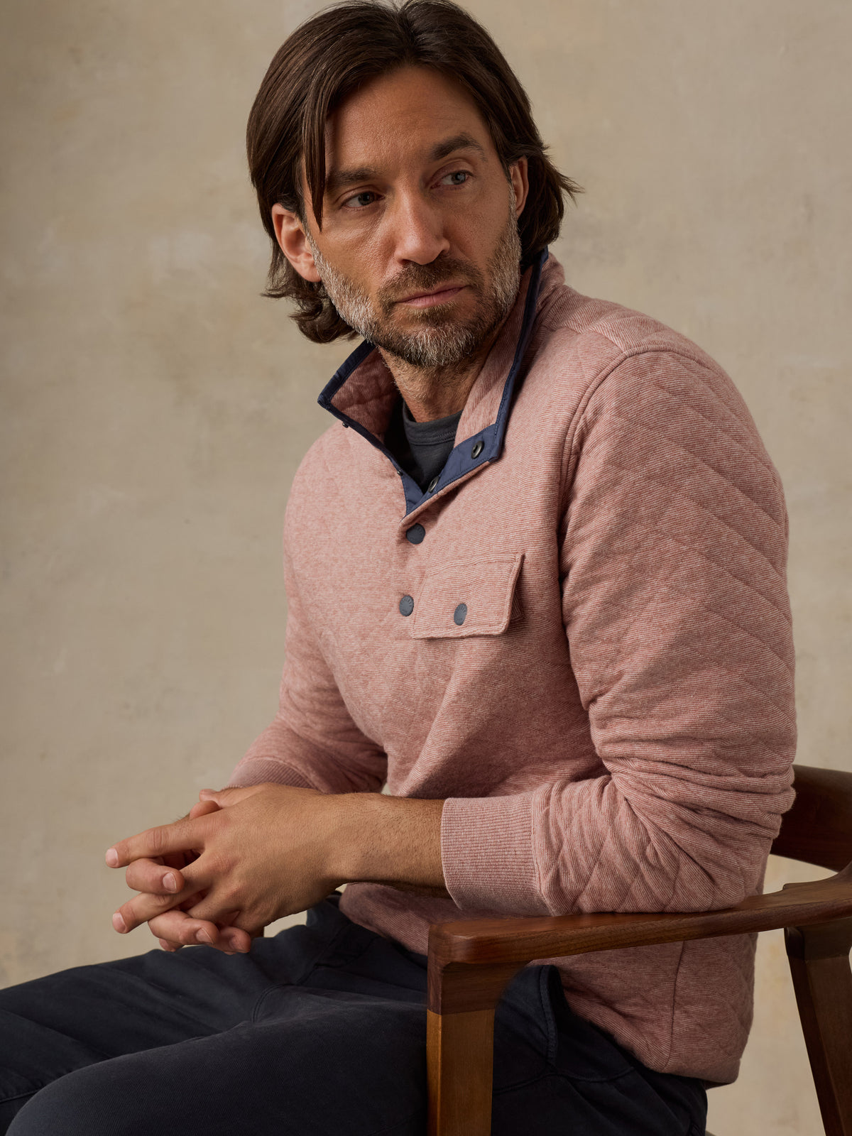 A man with shoulder-length brown hair and a beard sits in a wooden chair, wearing a dusty rose pink quilted long-sleeve henley sweater over a dark blue shirt and dark trousers, against a textured beige studio background., image 4 of 5