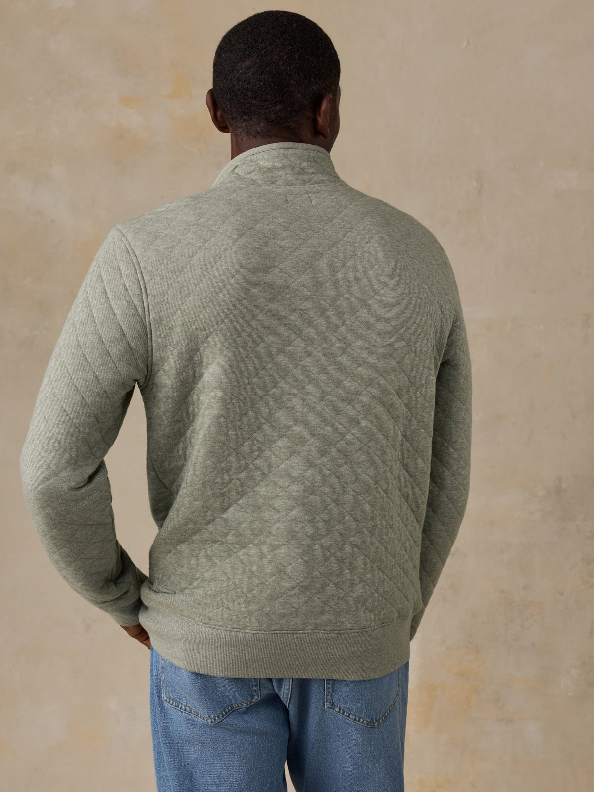 A man wearing a sage green quilted long-sleeve pullover and blue jeans, standing next to a wooden chair against a textured beige wall., image 3 of 6