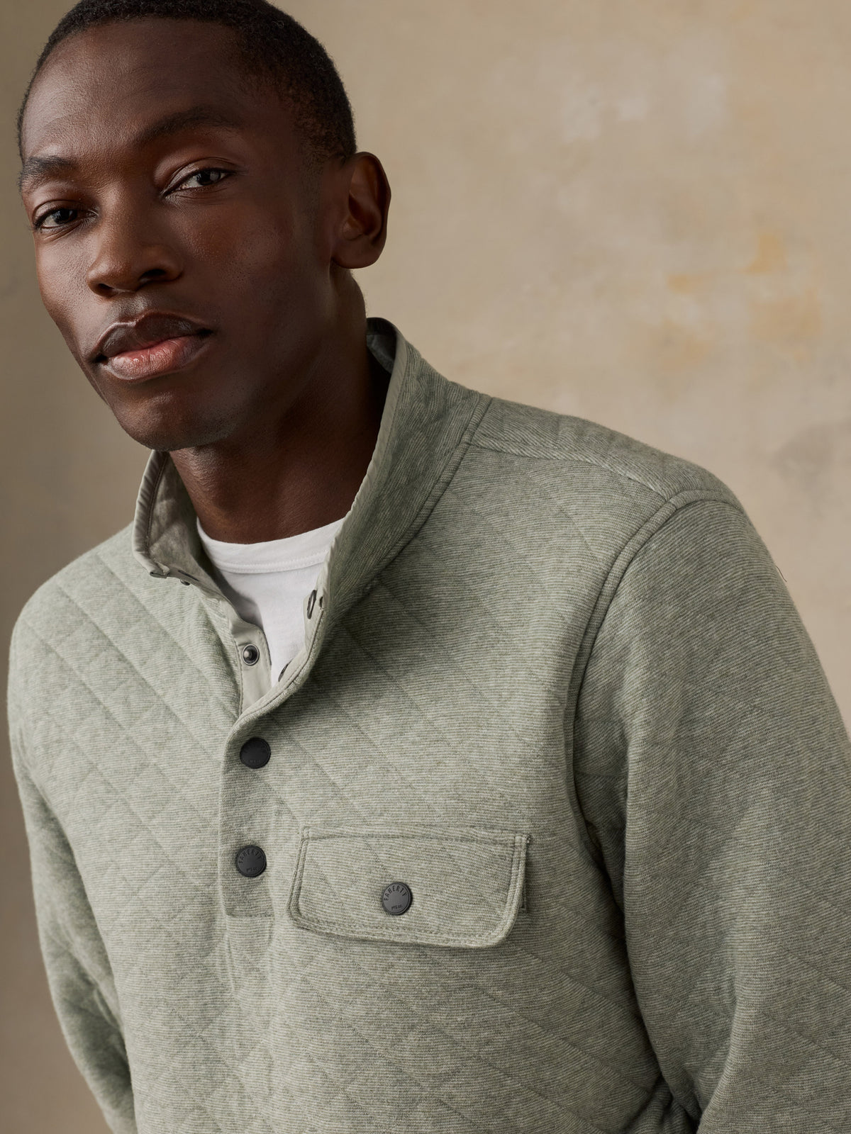 A man wearing a sage green quilted long-sleeve pullover over a white t-shirt, standing next to a wooden chair against a textured beige wall., image 2 of 6