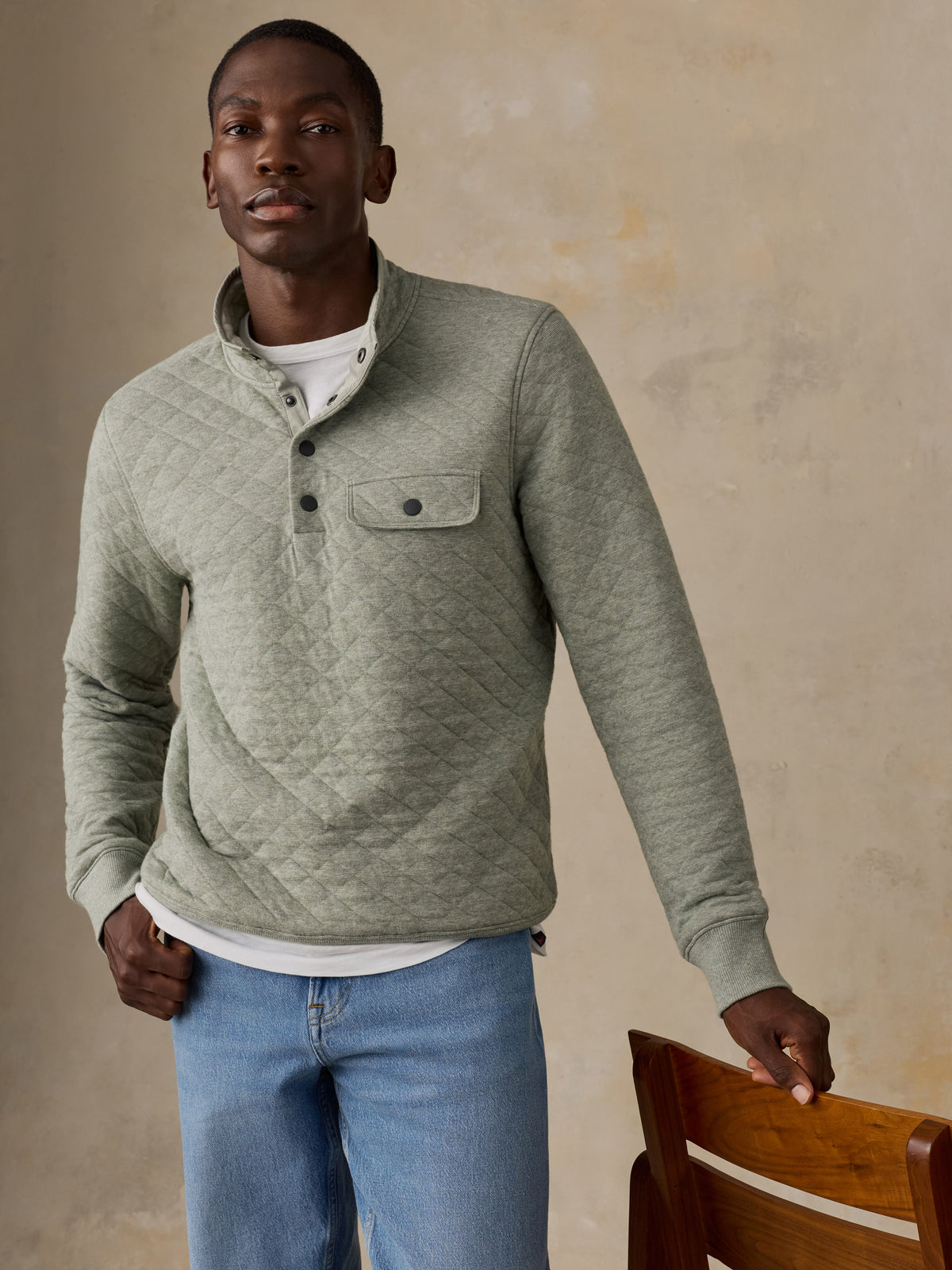 A man wearing a sage green quilted long-sleeve pullover over a white t-shirt and blue jeans, standing next to a wooden chair against a textured beige wall., image 1 of 6