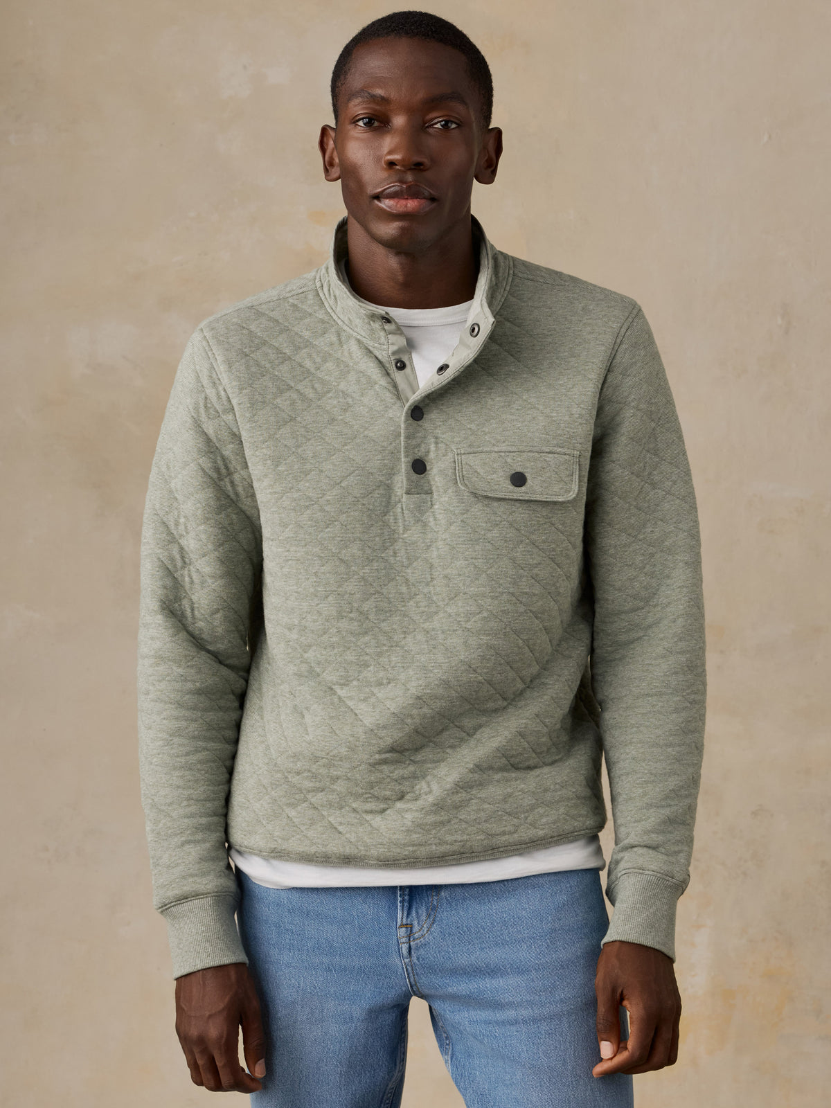 A man wearing a light sage green quilted long-sleeve pullover over a white t-shirt and blue jeans, standing against a textured beige background., image 6 of 6