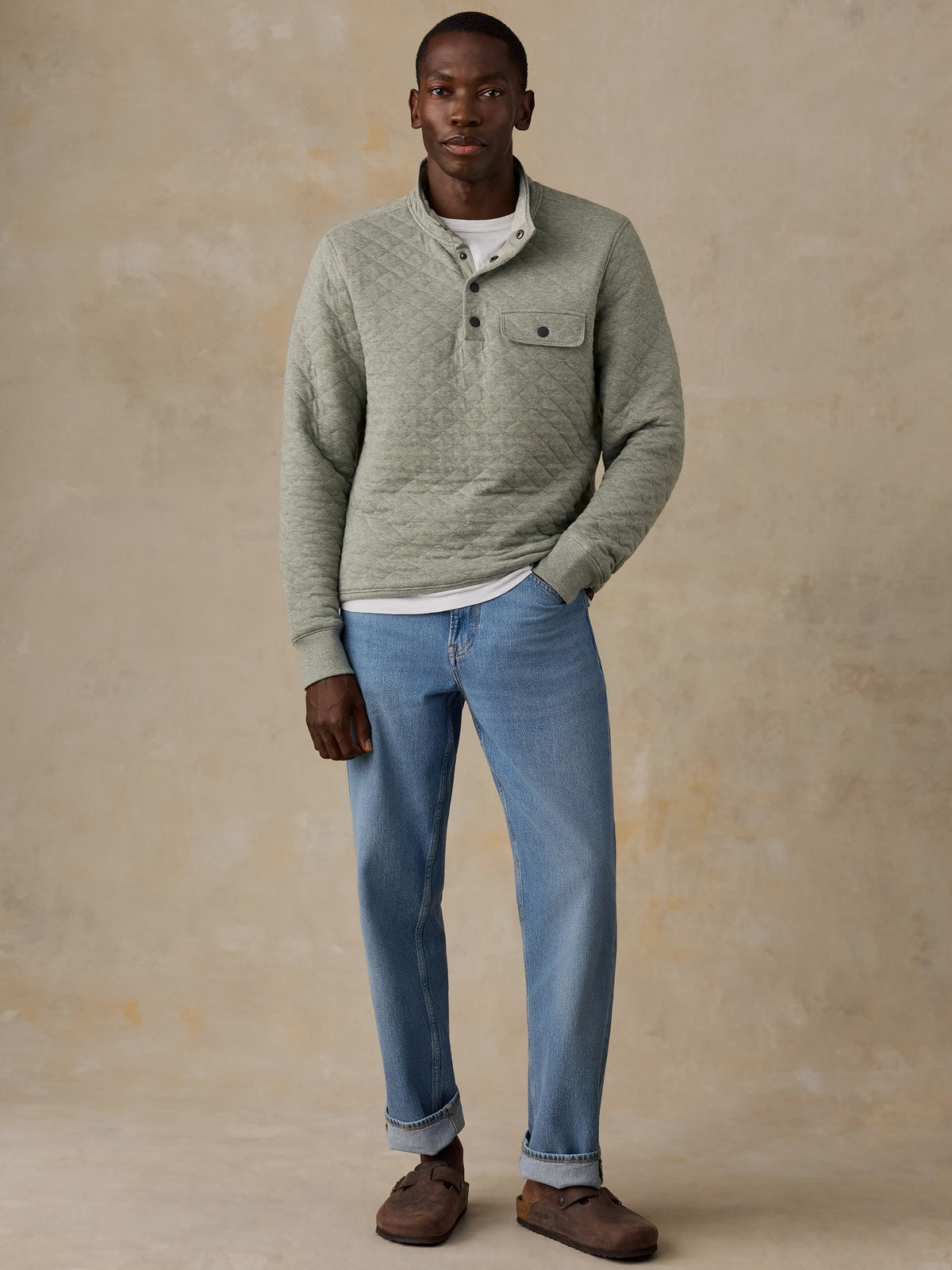 A man wearing a sage green quilted long-sleeve pullover over a white t-shirt and blue jeans, standing next to a wooden chair against a textured beige wall., image 4 of 6