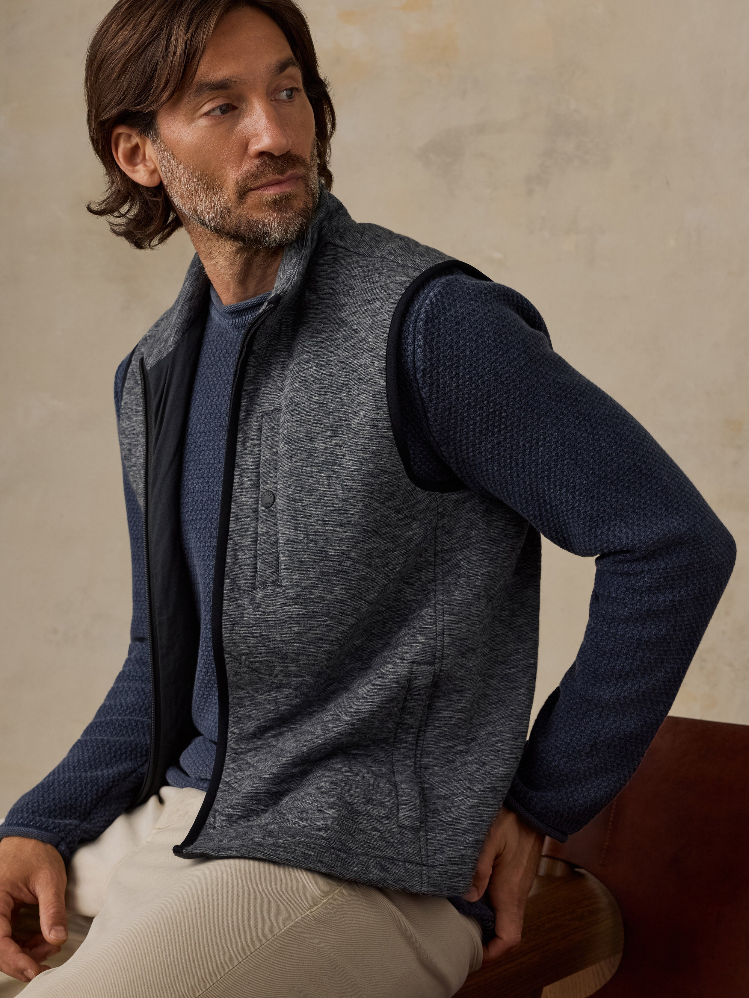 A man wearing a charcoal gray zip-front fleece vest over a navy blue textured knit sweater and light beige trousers, seated against a warm beige textured wall.