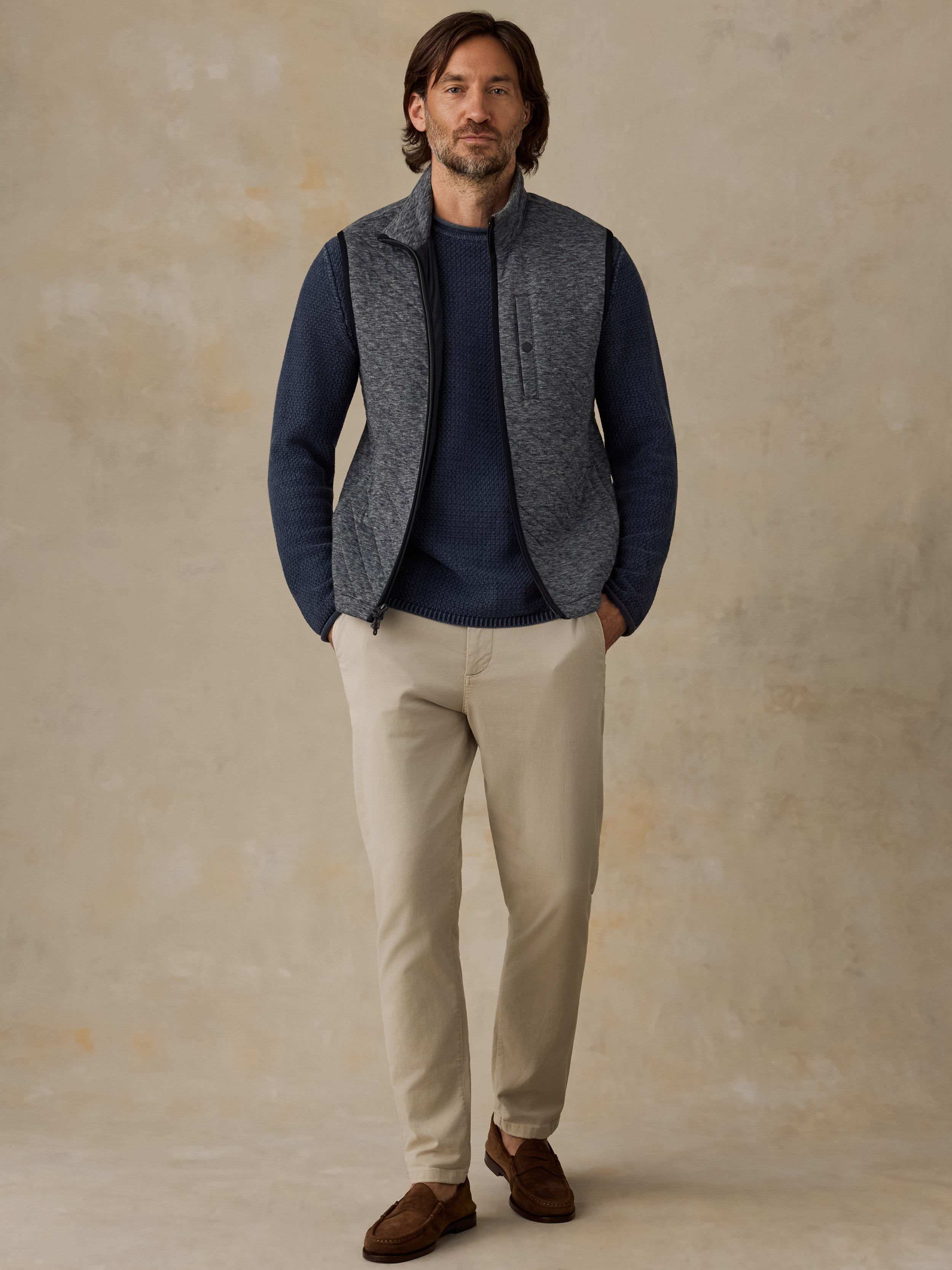 A man wearing a gray heathered zip-front fleece vest over a navy blue sweater, paired with beige trousers and brown shoes, standing against a textured beige studio backdrop.