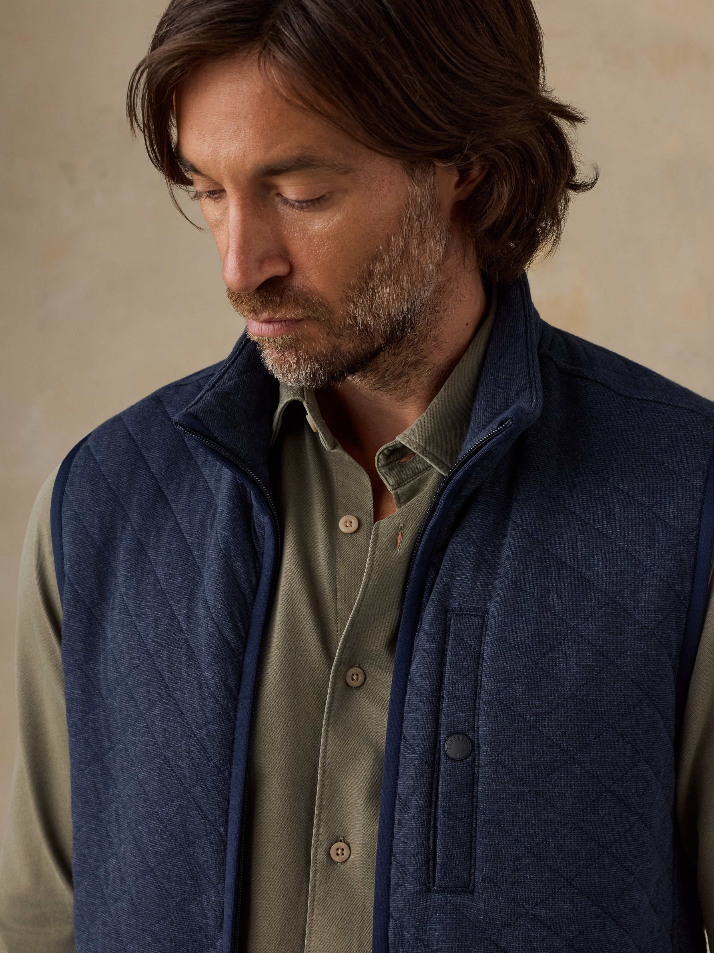 A man wearing a navy blue quilted vest over an olive green button-down shirt, shown in a close-up portrait against a neutral beige background.