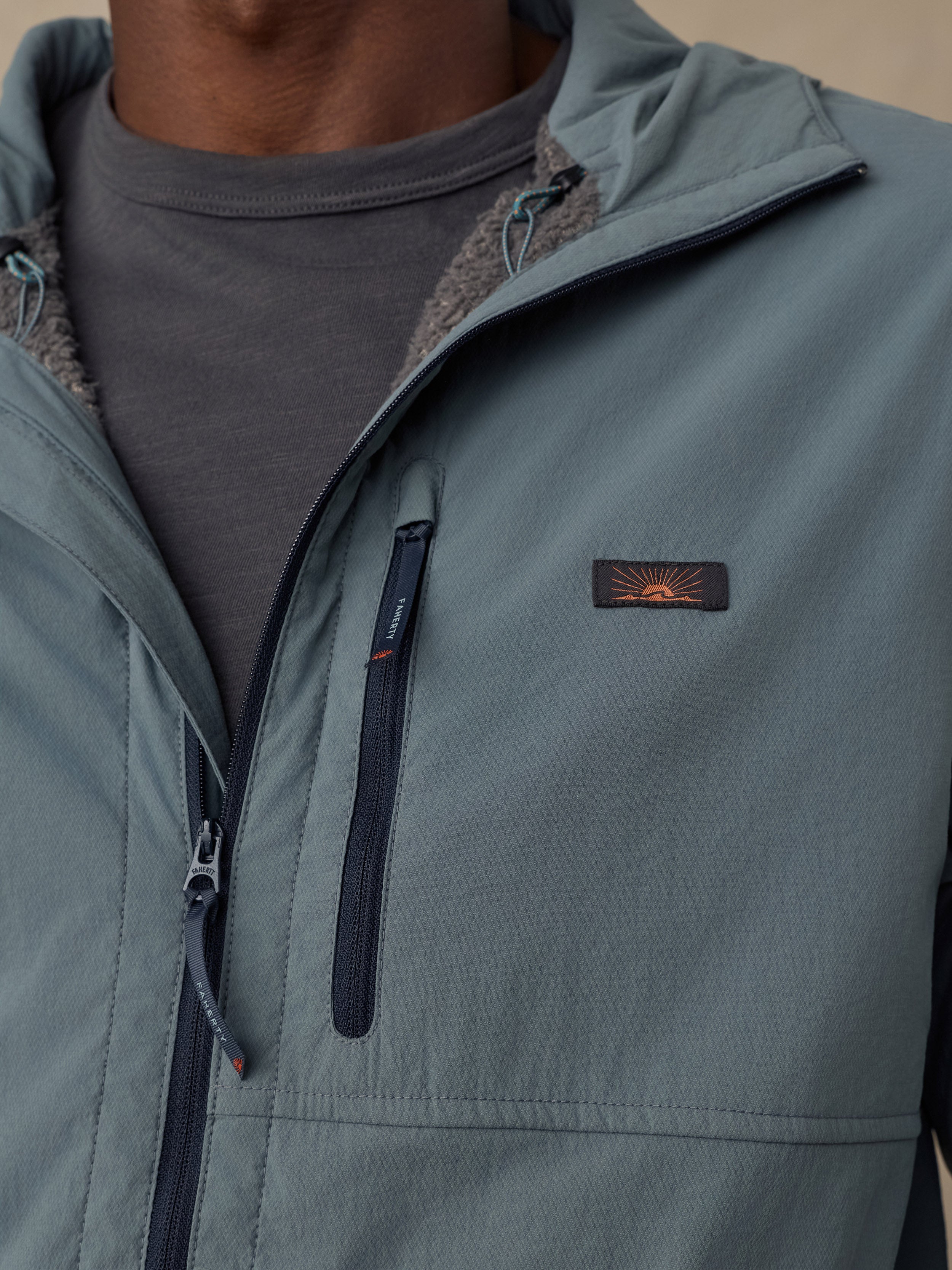 Close-up of a man wearing a muted blue-gray zip-front jacket over a dark gray shirt. The jacket has a Faherty sun and waves chest logo and a vertical zip pocket with a black zipper pull.
