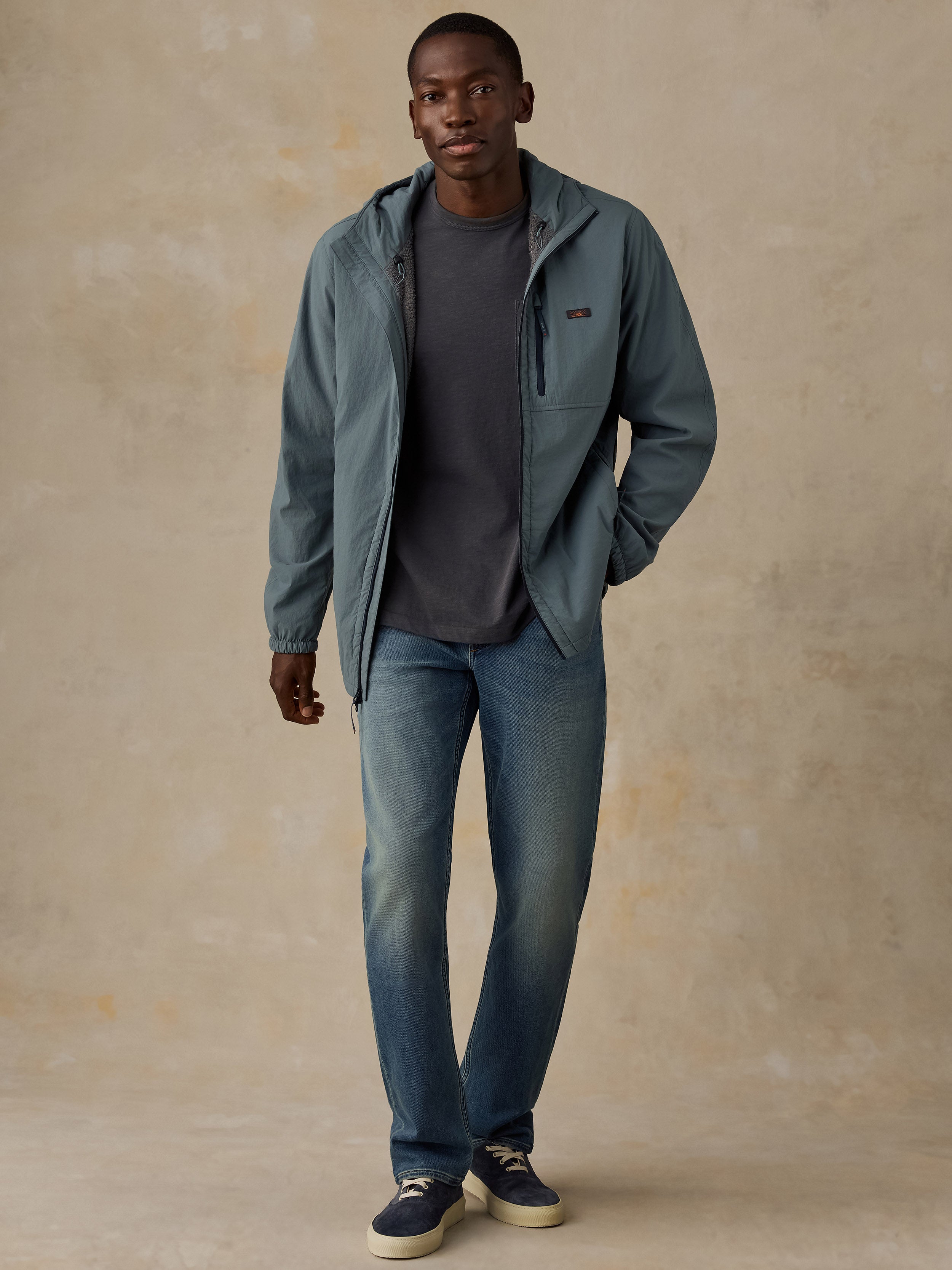 A man modeling a slate blue zip-up hooded jacket over a dark gray t-shirt and blue jeans, standing against a textured beige studio backdrop.