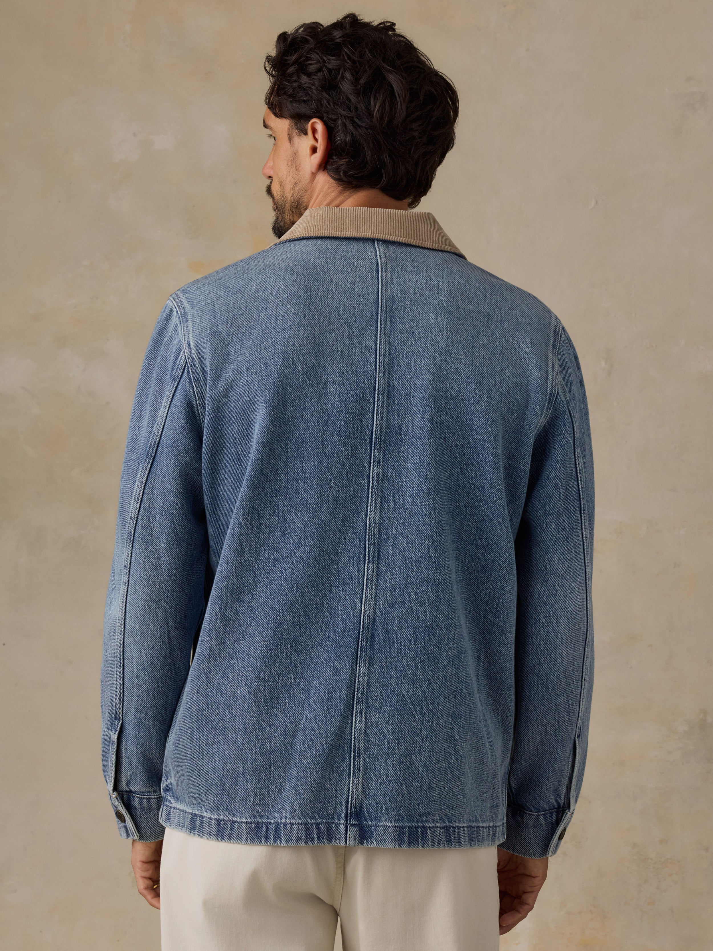 Man wearing a blue denim jacket with tan corduroy collar and white chino pants against a beige background