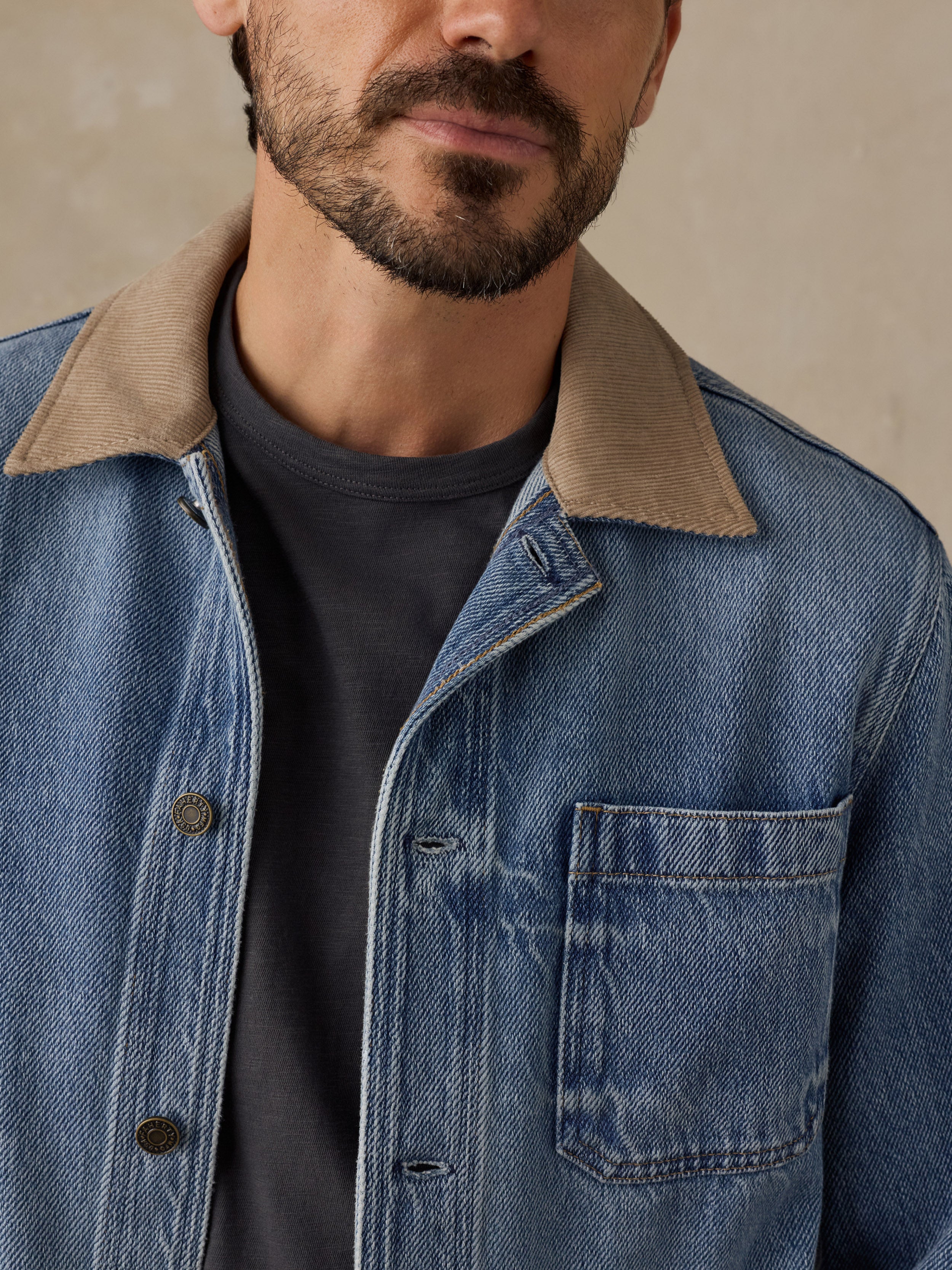Man wearing a blue denim jacket with tan corduroy collar and a blue t-shirt against a beige background