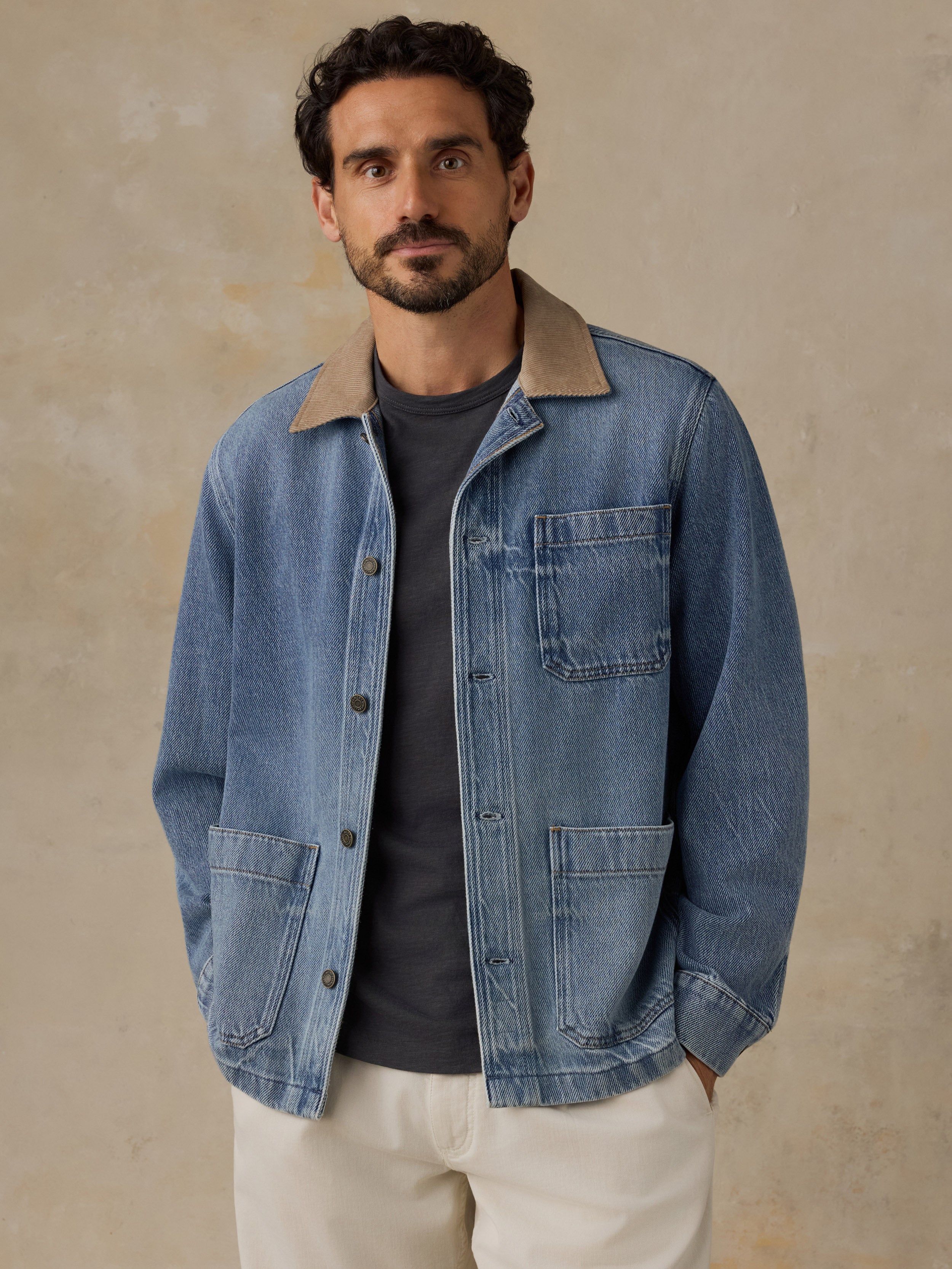 Man wearing a blue denim jacket with tan corduroy collar, blue t-shirt, and white chino pants against a beige background