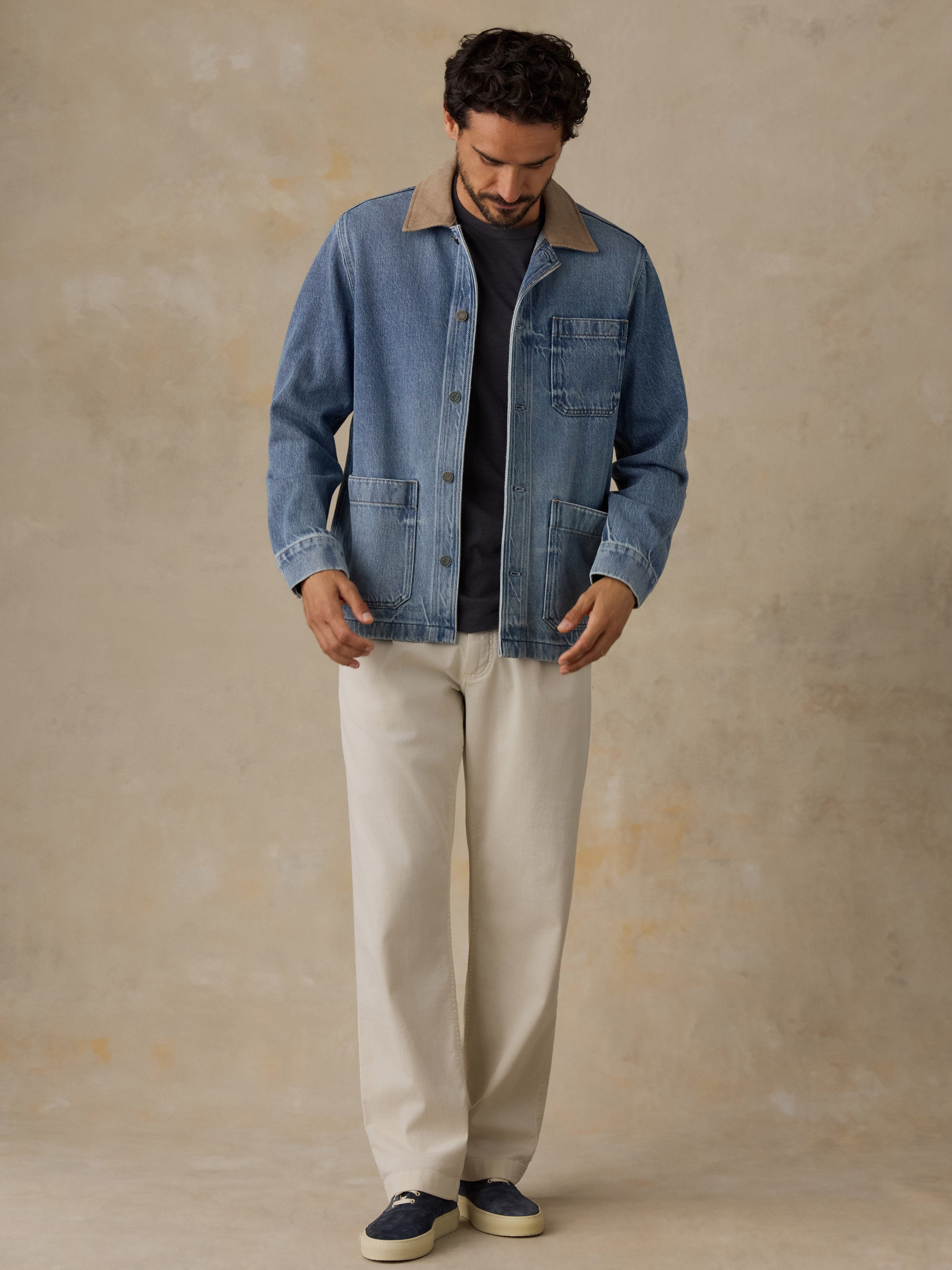 Man wearing a blue denim jacket with tan corduroy collar, blue t-shirt, and white chino pants against a beige background