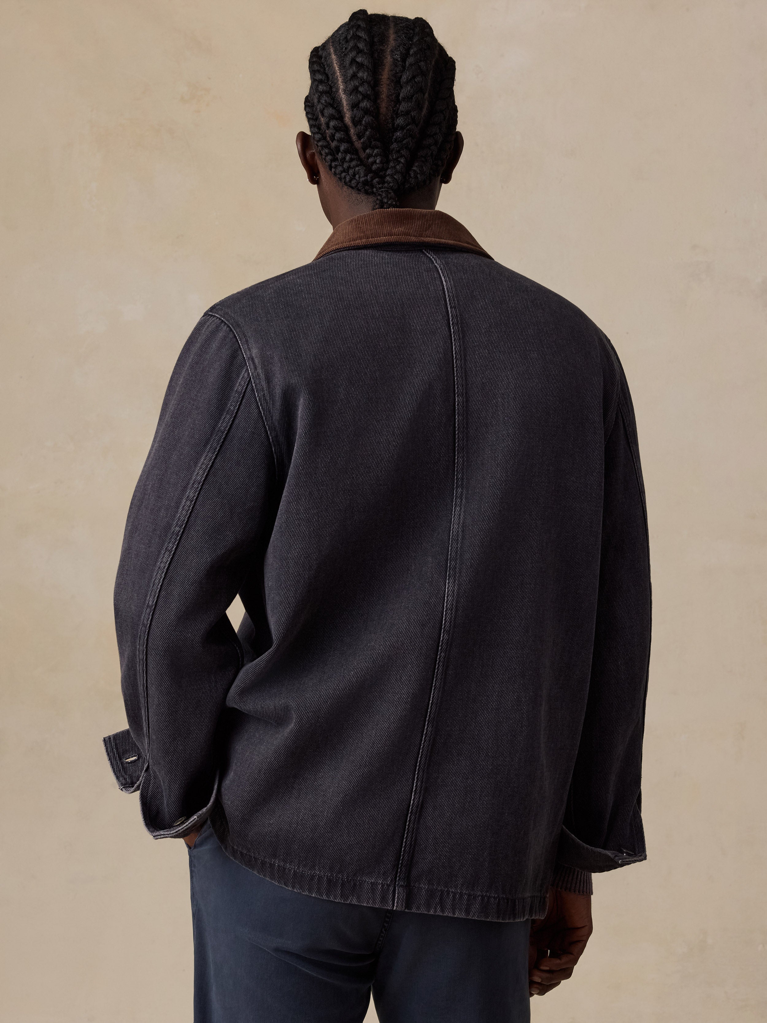 A man viewed from behind wearing a dark charcoal gray canvas jacket with a brown corduroy collar, set against a textured beige background.