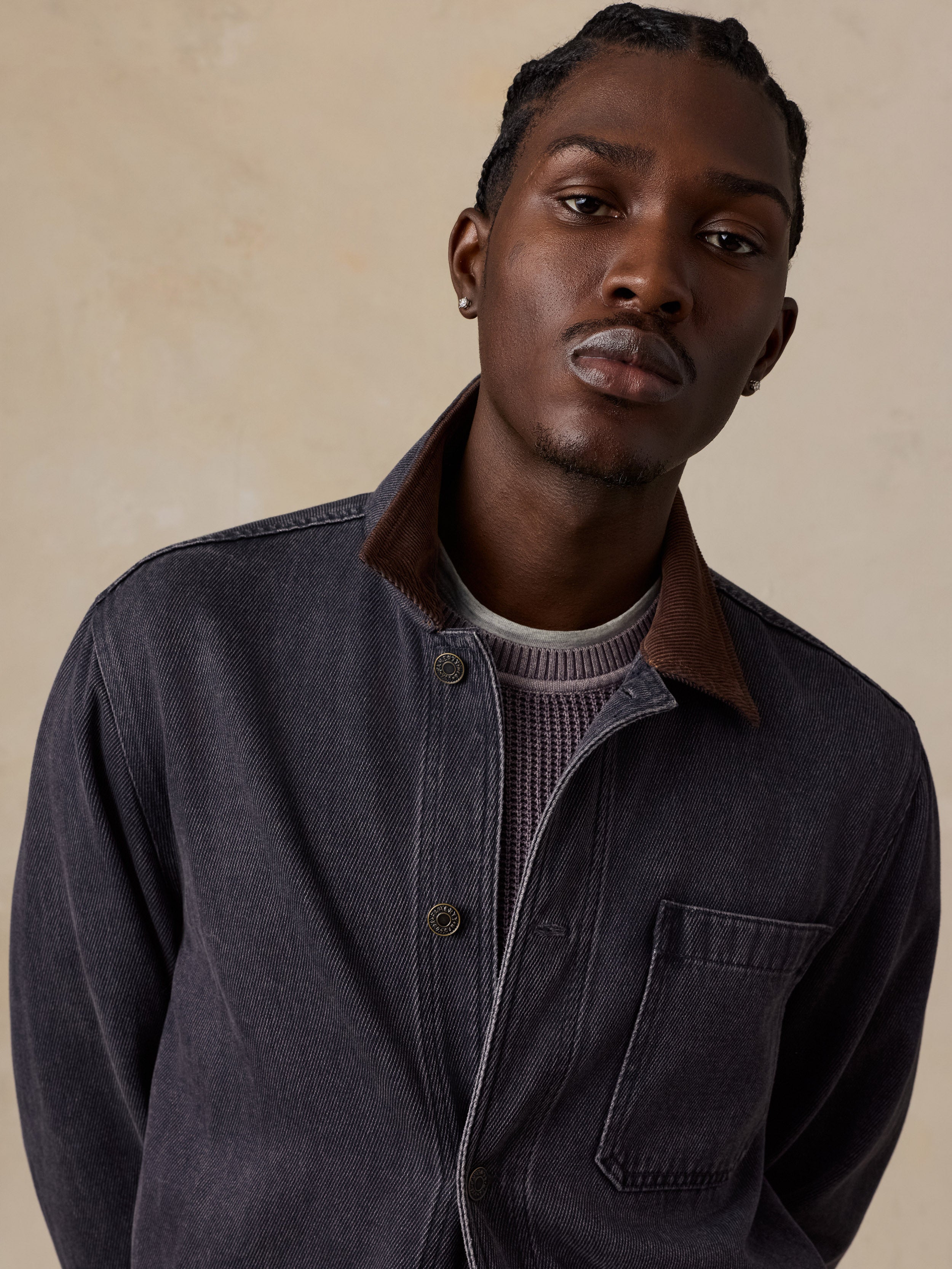 A man wearing a dark charcoal gray corduroy overshirt with a brown corduroy collar over a textured gray knit sweater, set against a warm beige background.