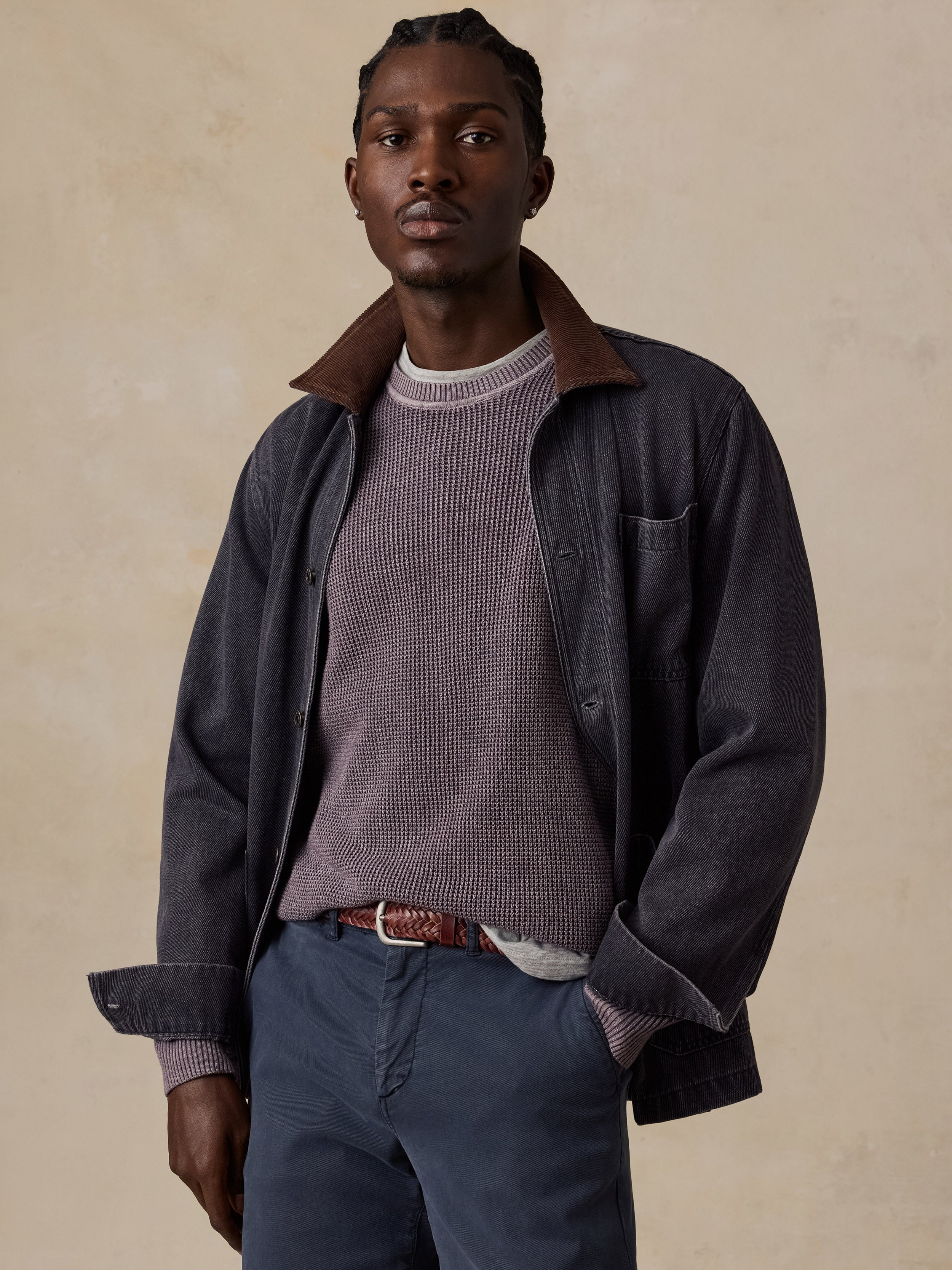 A man wearing a dark charcoal corduroy overshirt with a brown collar, layered over a textured mauve knit sweater, blue trousers, and a brown belt, posed against a warm beige textured studio backdrop.