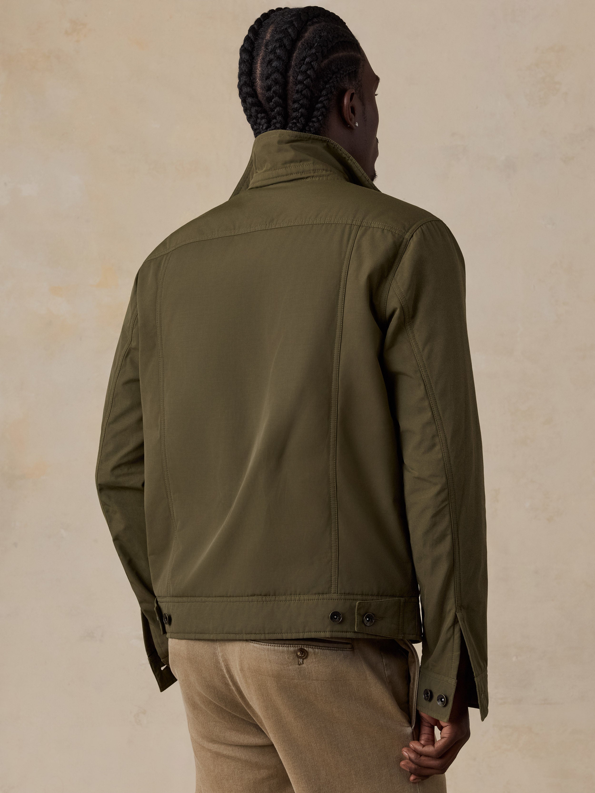 A man shown from the back wearing an olive green utility jacket and beige trousers, standing against a textured beige studio background.